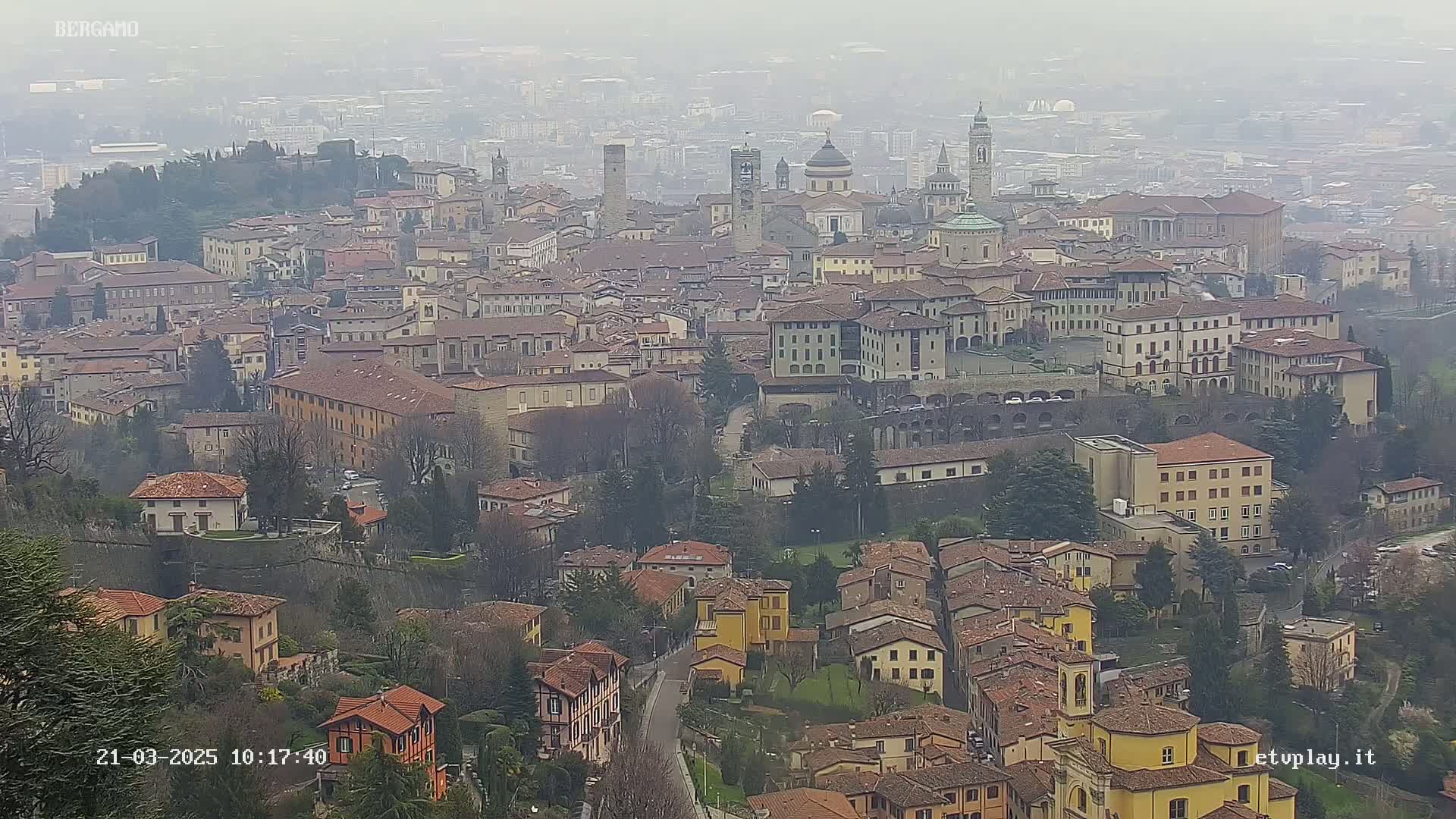 A hazy, high-angle view shows a sprawling city of many low-rise buildings with red-tiled roofs, interspersed with taller structures and several prominent churches, all set amongst bare winter trees.