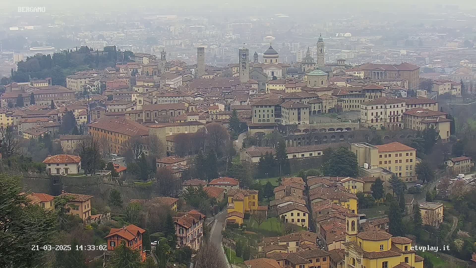 A hazy view of a dense European city, featuring numerous buildings with red tile roofs, situated on a hillside.