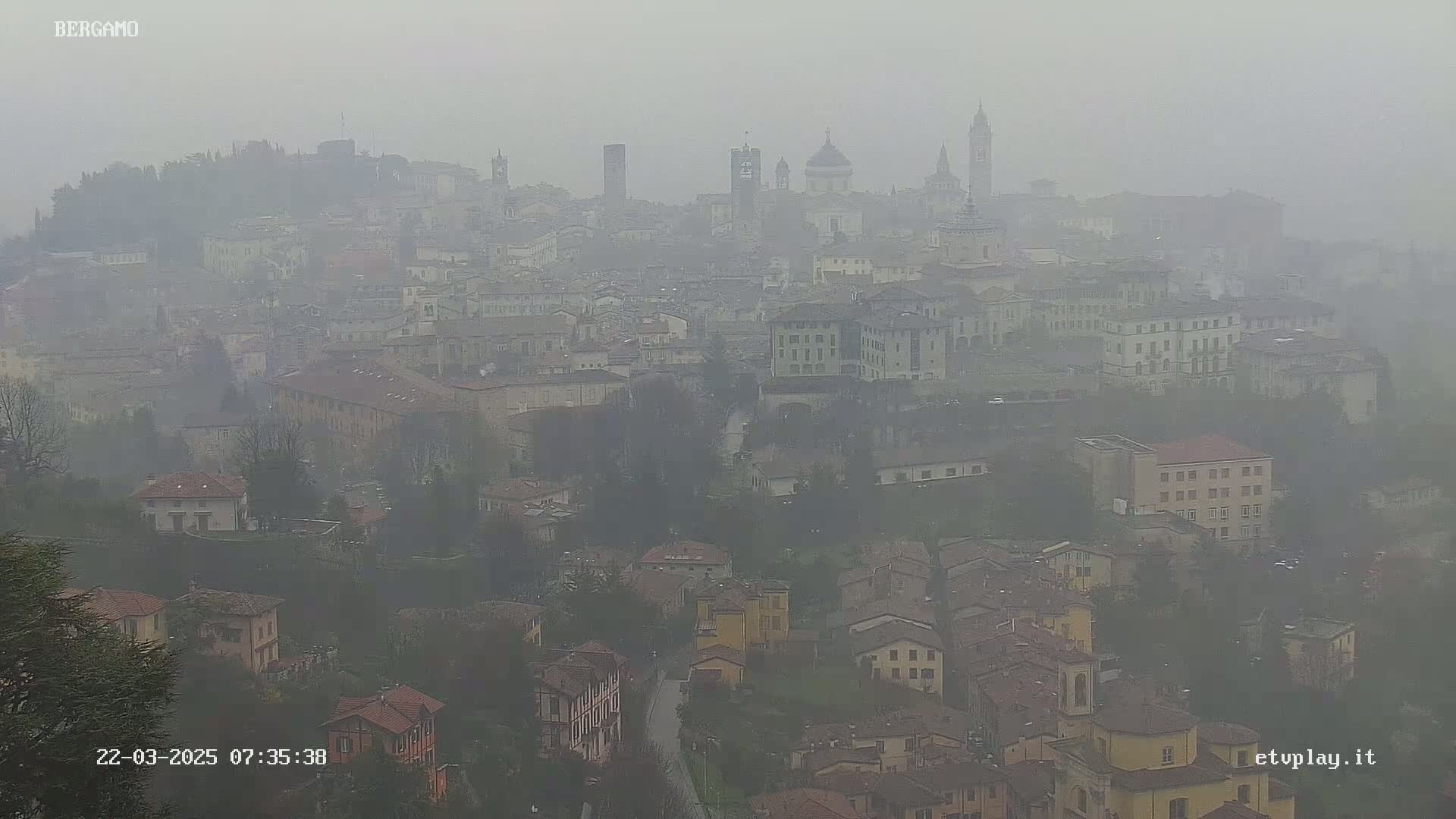 A hazy, foggy view of a city with numerous buildings, including several prominent towers and domes, stretches out across a hillside.