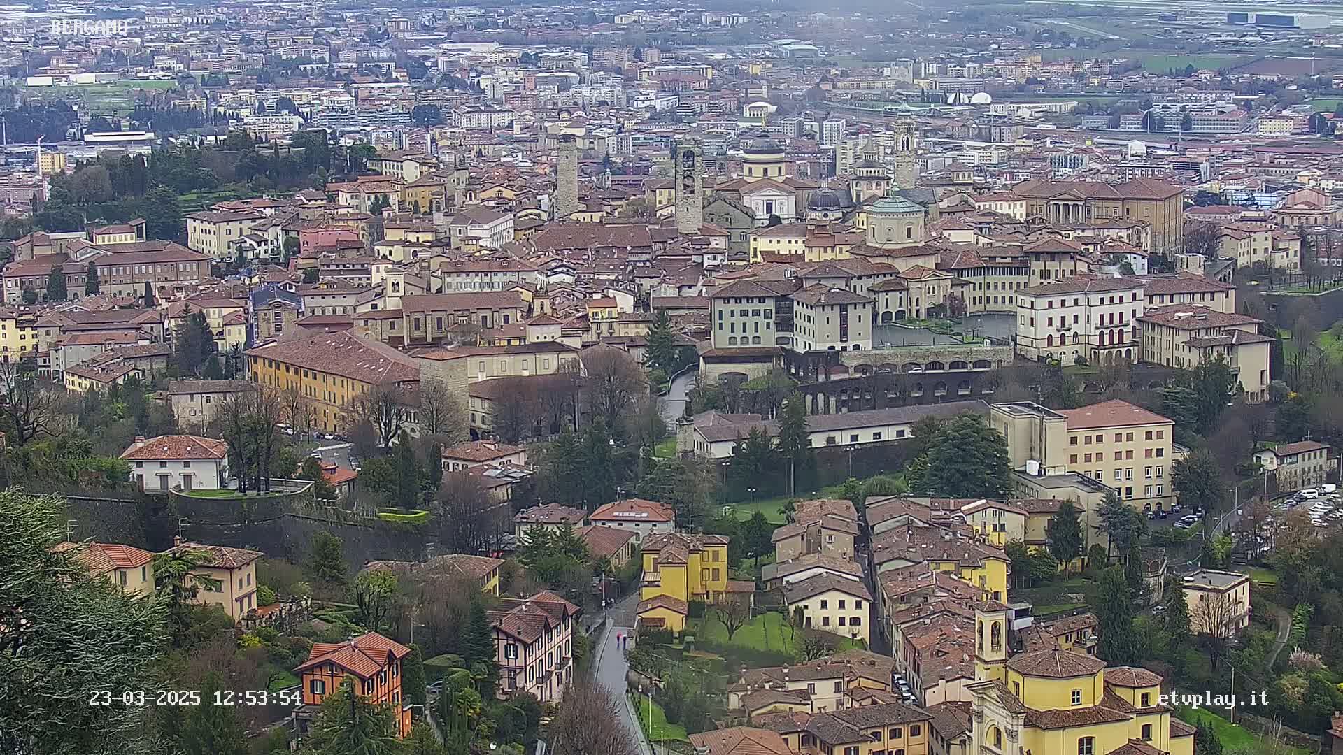 An aerial view of a dense, historic city with many brown-roofed buildings, under an overcast sky.