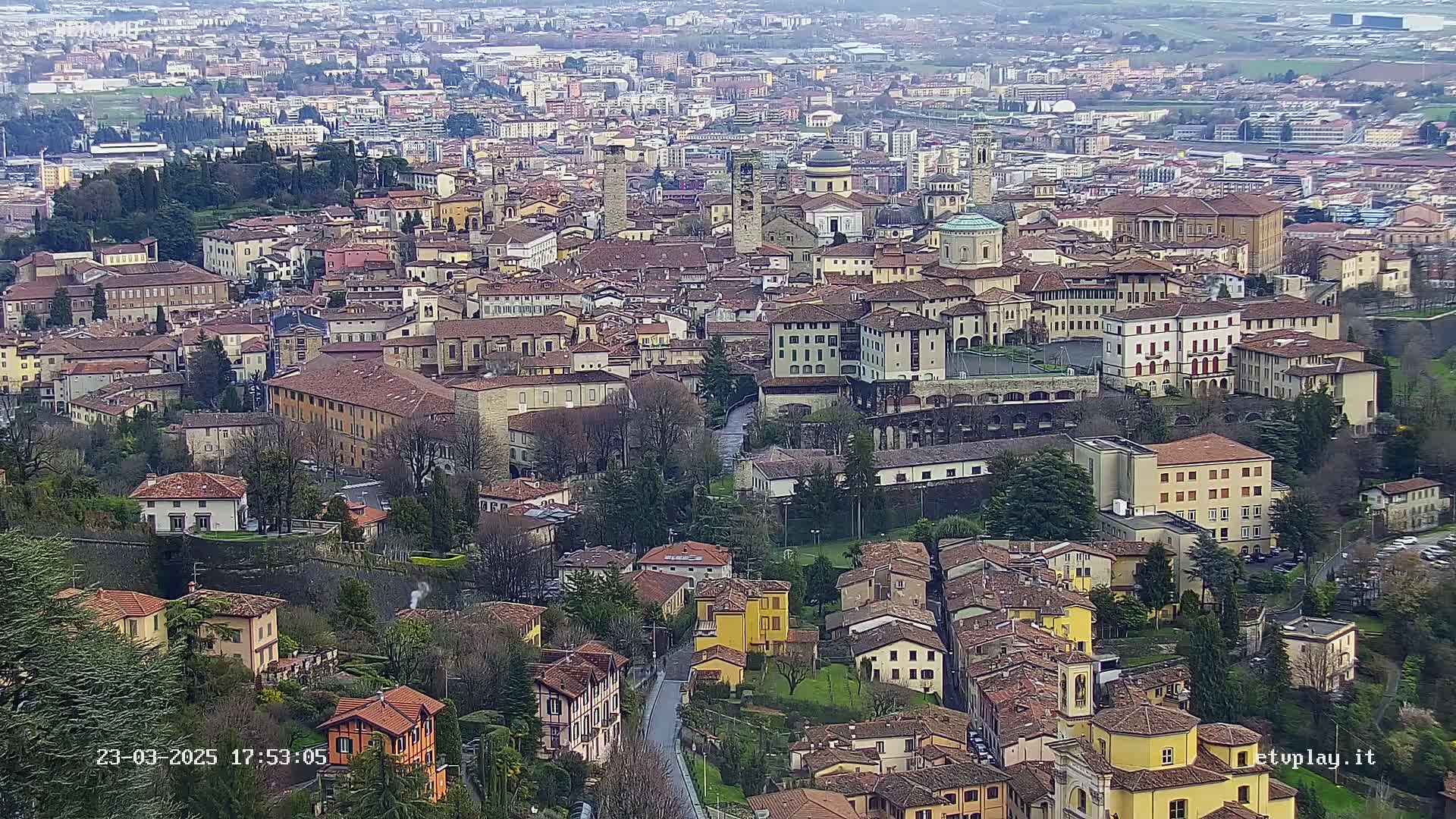 An aerial view shows a sprawling city with numerous terracotta-roofed buildings nestled amongst green spaces, under an overcast sky.