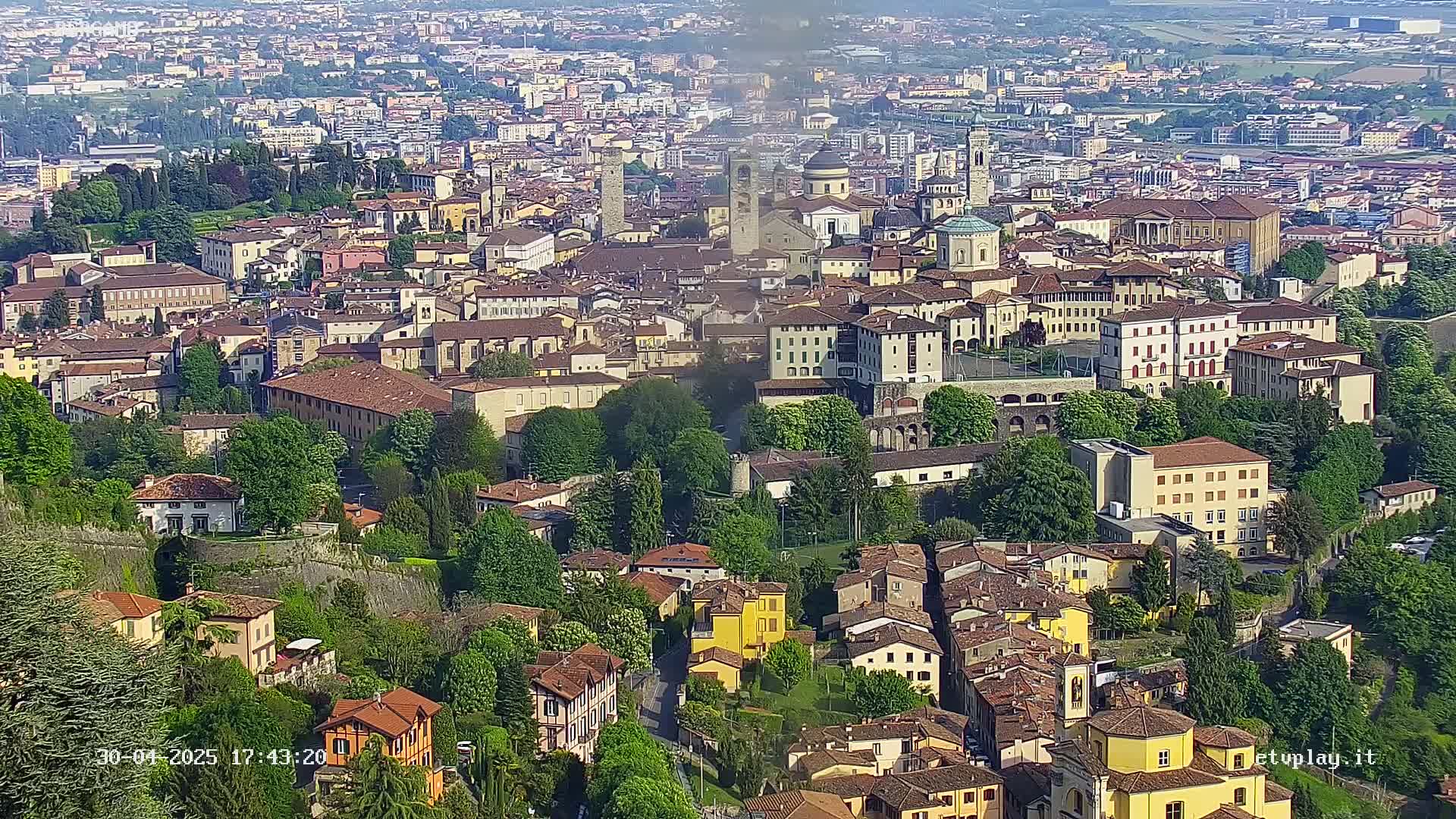 A sprawling, historic city with numerous buildings of varying heights and styles is nestled amongst lush green trees and hills on a partly sunny day.
