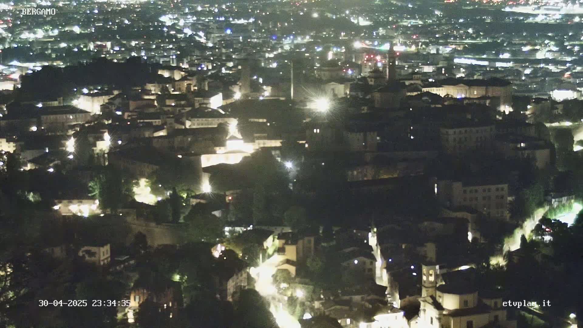 A nighttime, aerial view of a city with numerous lights illuminating buildings and streets nestled among dark green areas.
