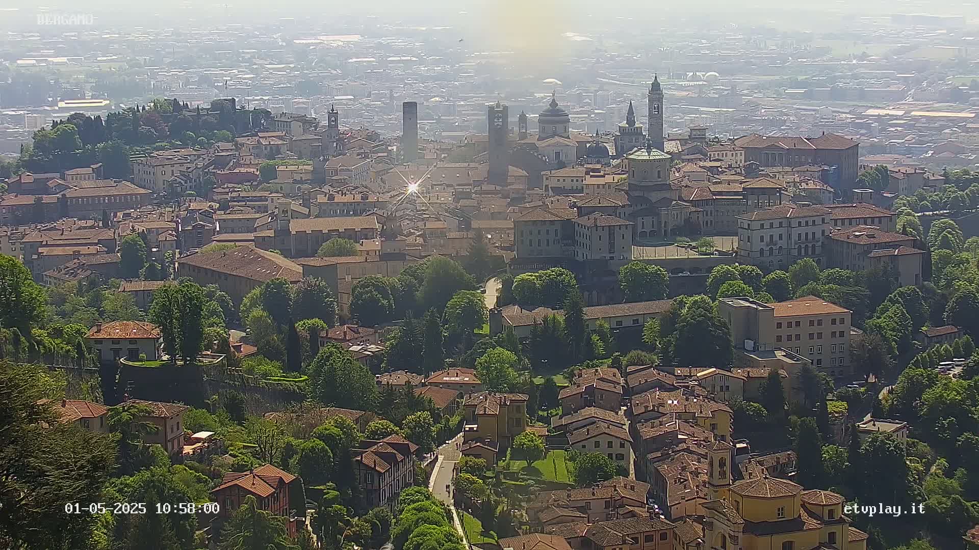 A hazy, sunny day reveals a sprawling city of terracotta-roofed buildings, interspersed with lush green trees and punctuated by several prominent church steeples and towers.
