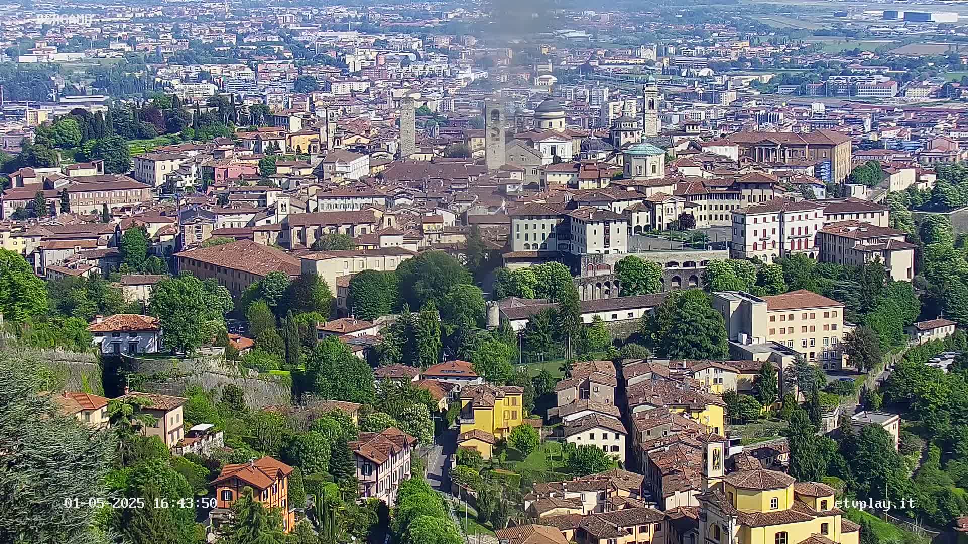 A sprawling city with red-tiled roofs and green spaces, viewed from above on a clear, sunny day.