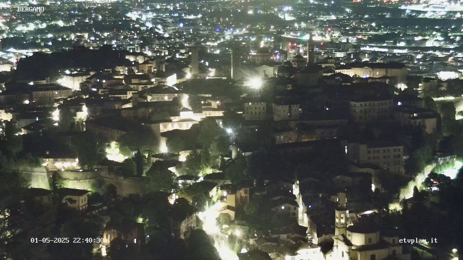 A nighttime aerial view shows a city illuminated by lights, nestled among dark green trees and hills.