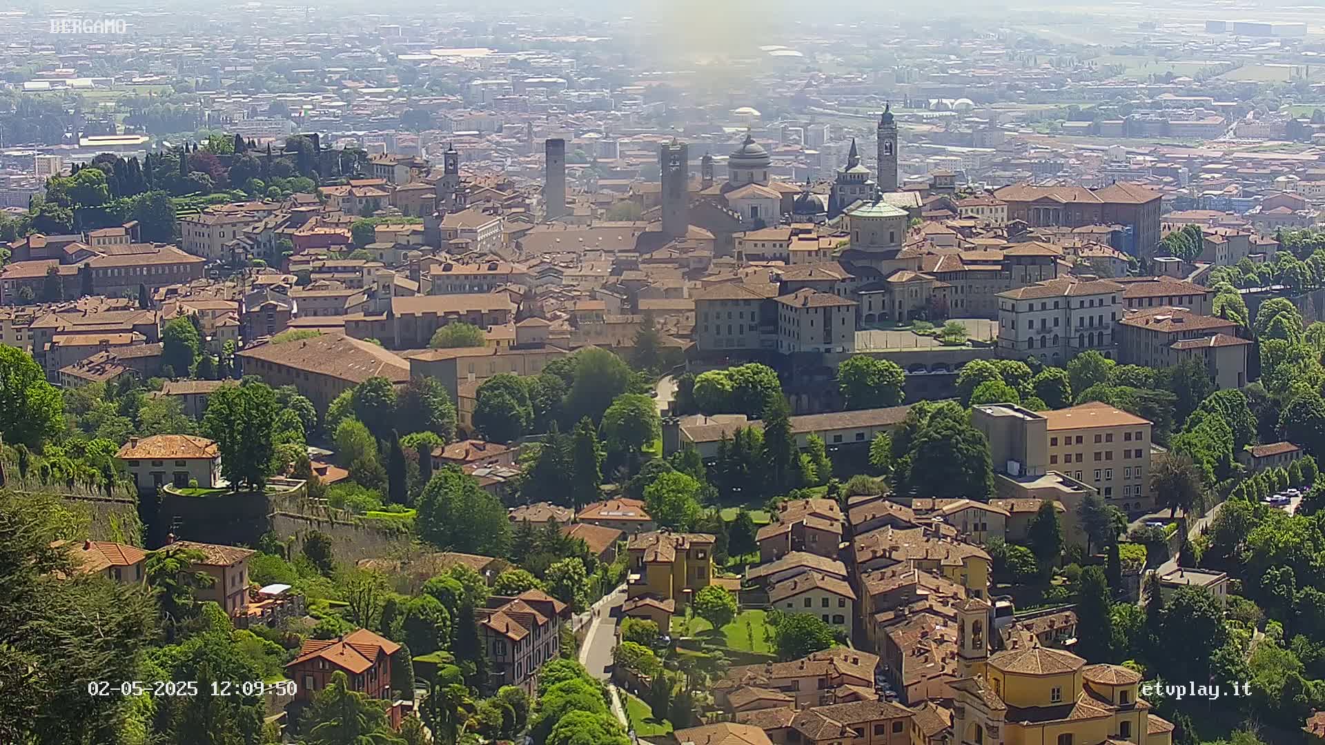 A sprawling city of red-roofed buildings nestled among lush green trees and hills under a hazy, sunny sky.