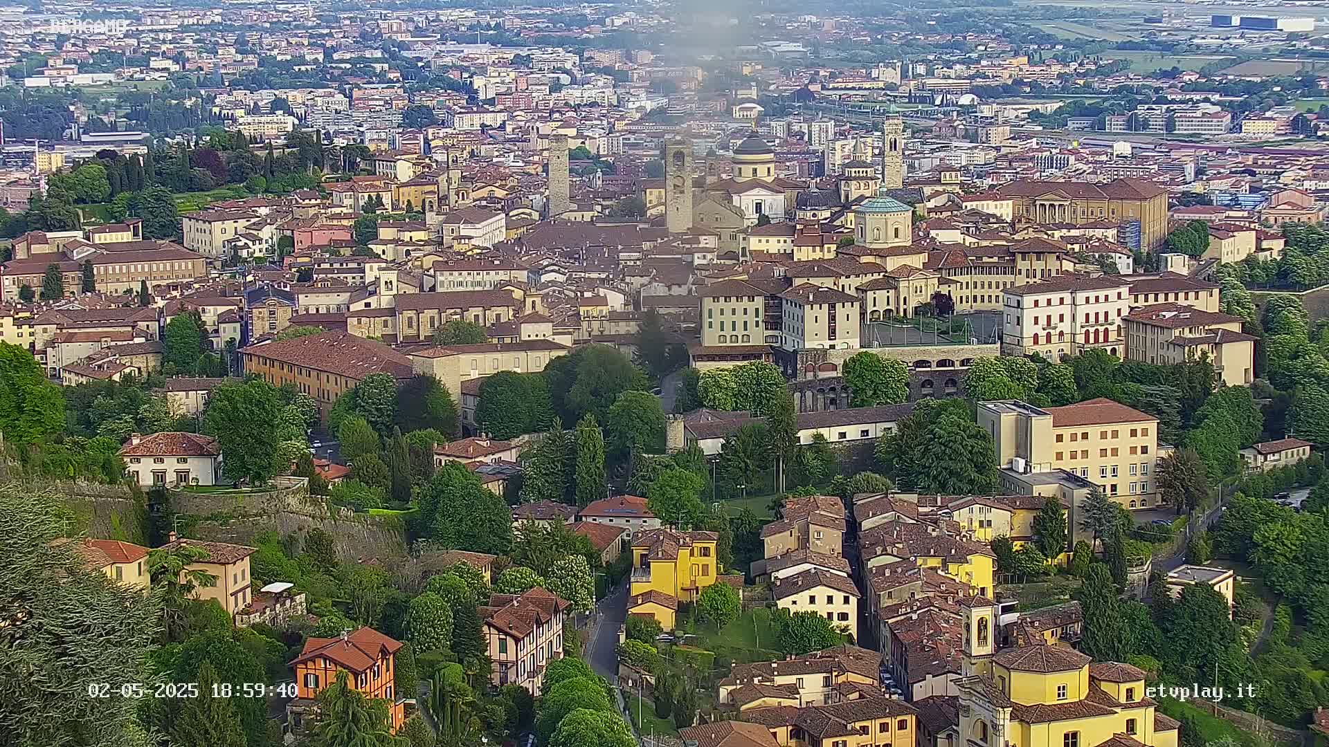 An aerial view shows a densely packed city with numerous brown-roofed buildings, surrounded by lush green trees and walls, under a partly cloudy sky.
