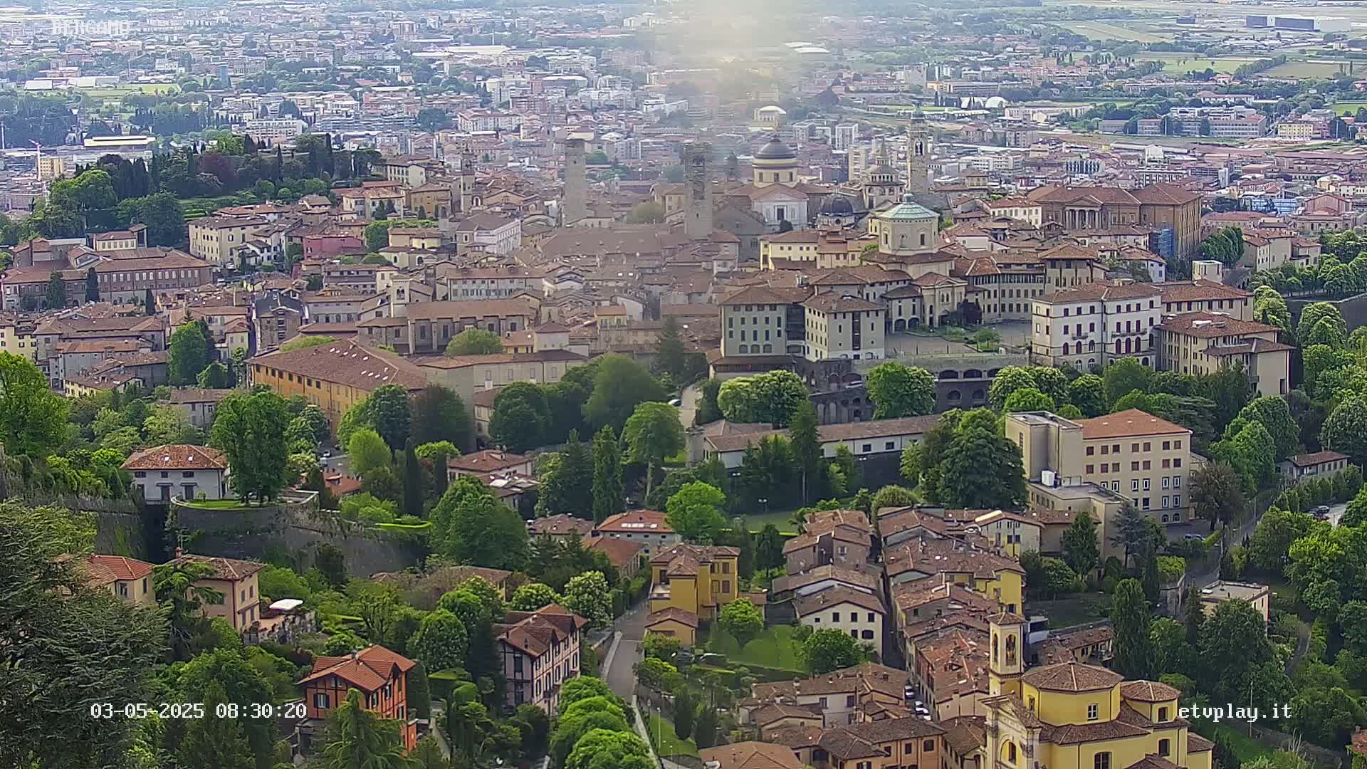 An aerial view shows a densely packed city of tan and brown buildings surrounded by lush green trees under a partly sunny sky.