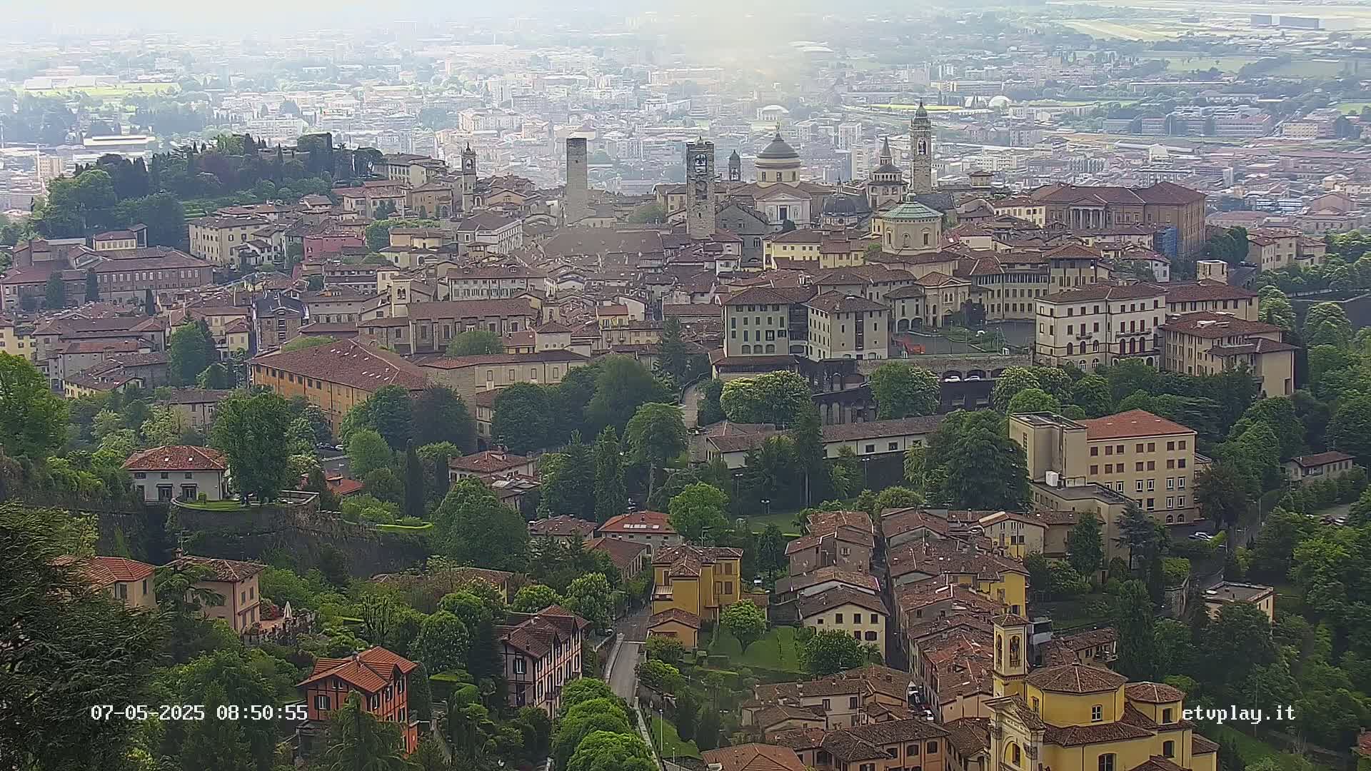 A hazy, aerial view shows a sprawling city with numerous buildings of varying heights and styles nestled amongst lush green trees.
