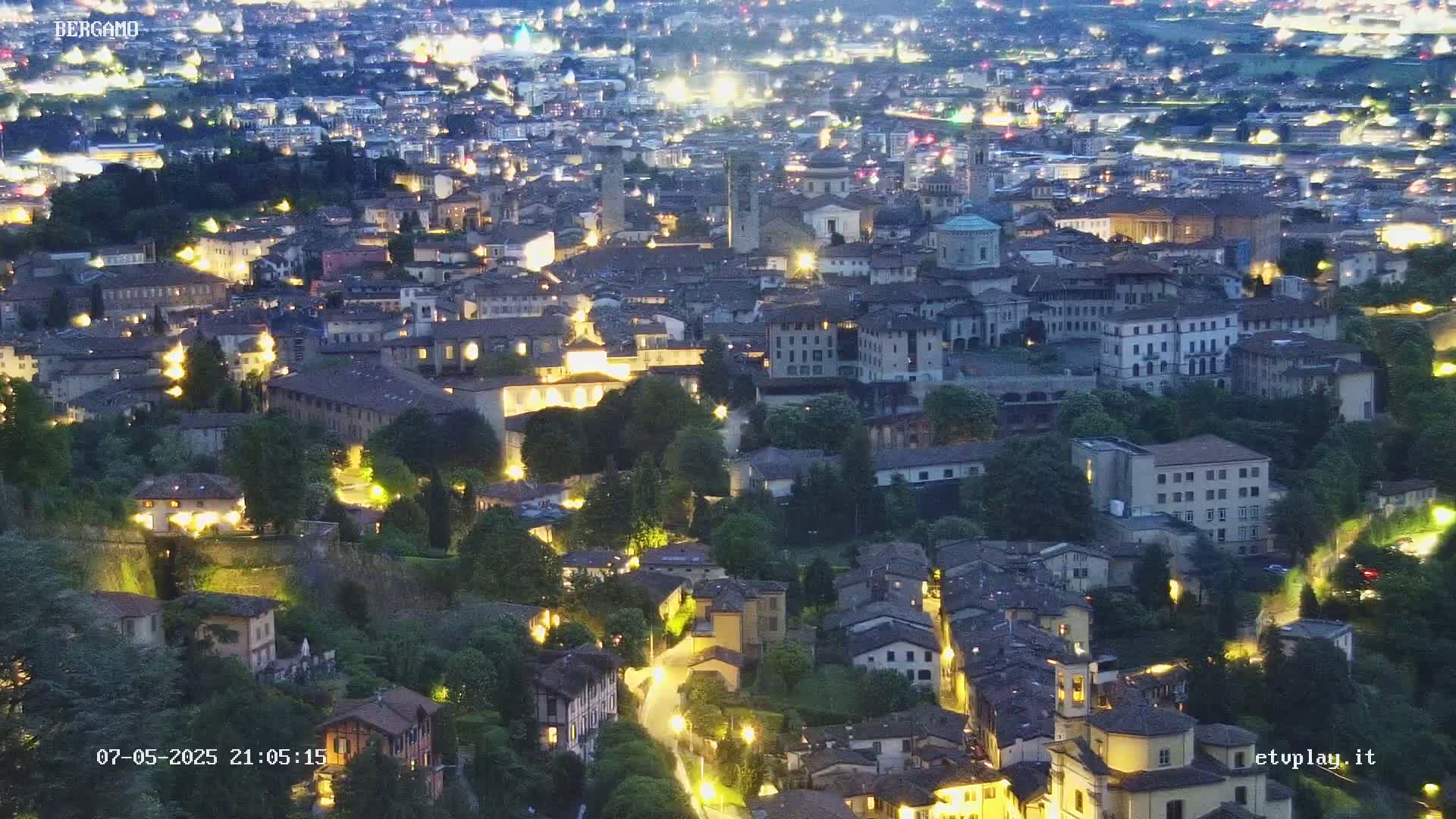A sprawling city at night, illuminated by numerous lights, nestled among green hills under a dark sky.