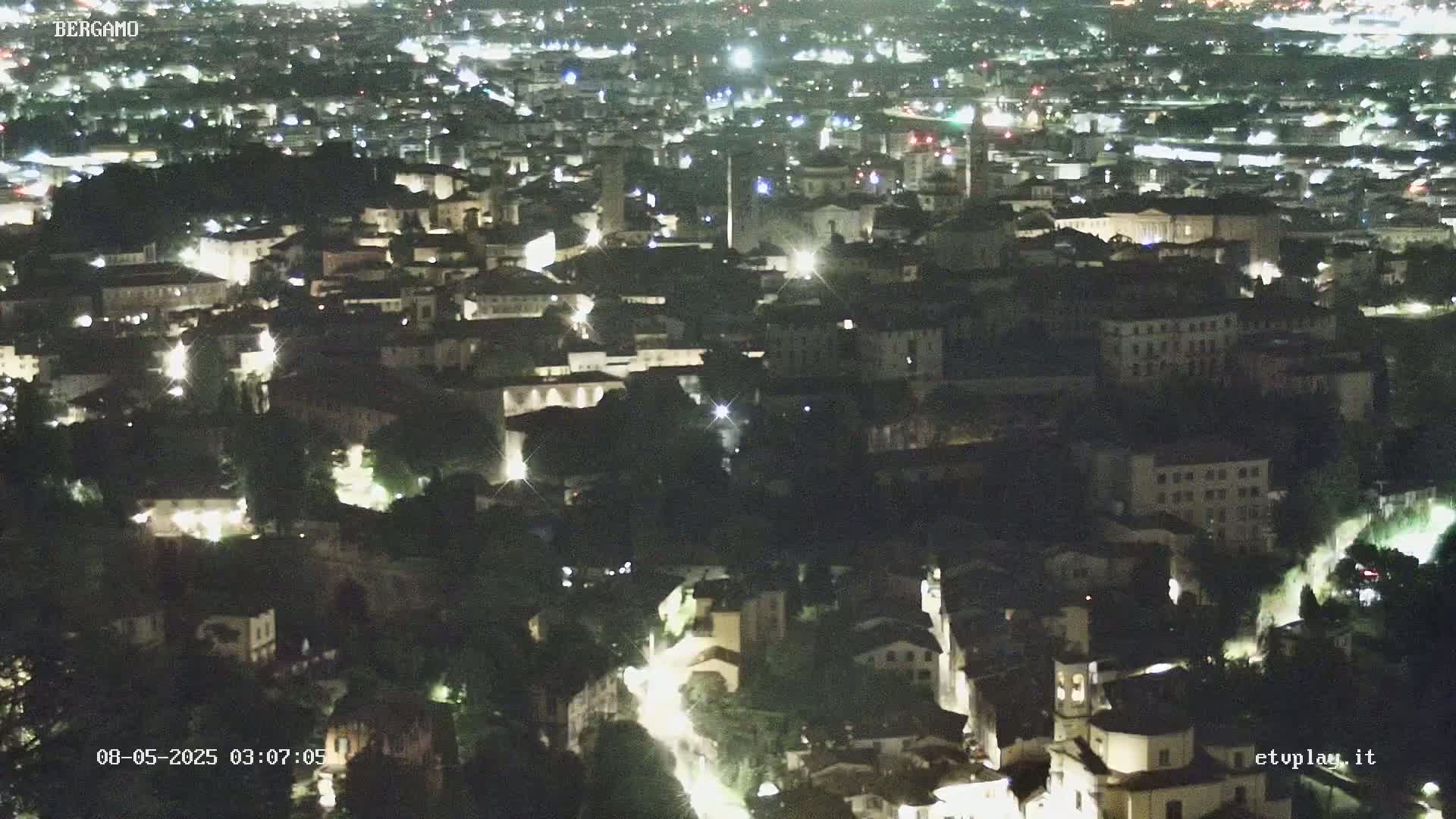 A nighttime, aerial view of a city with many buildings illuminated, nestled amongst dark, tree-covered areas.