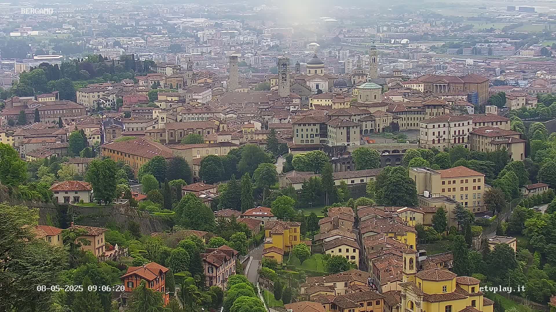 A hazy, high-angle view shows a large city nestled in a valley, filled with closely packed buildings of varying heights and styles, surrounded by lush green trees and walls.