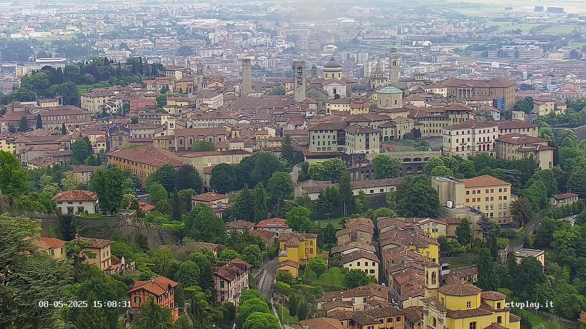 A sprawling city with numerous red-roofed buildings and abundant green spaces is viewed from above on an overcast day.