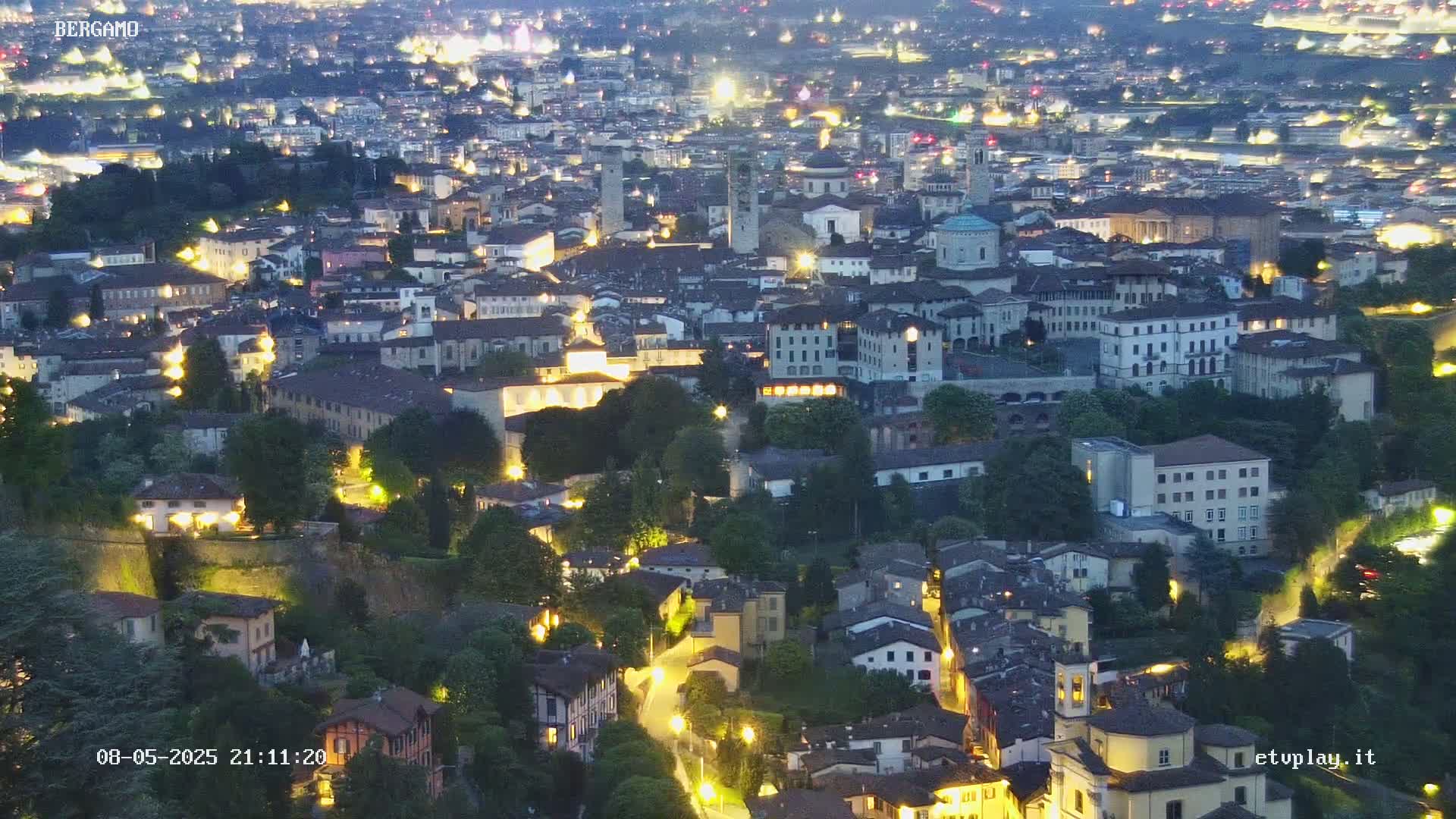 A nighttime aerial view of a city, illuminated by many lights, nestled among green hills.