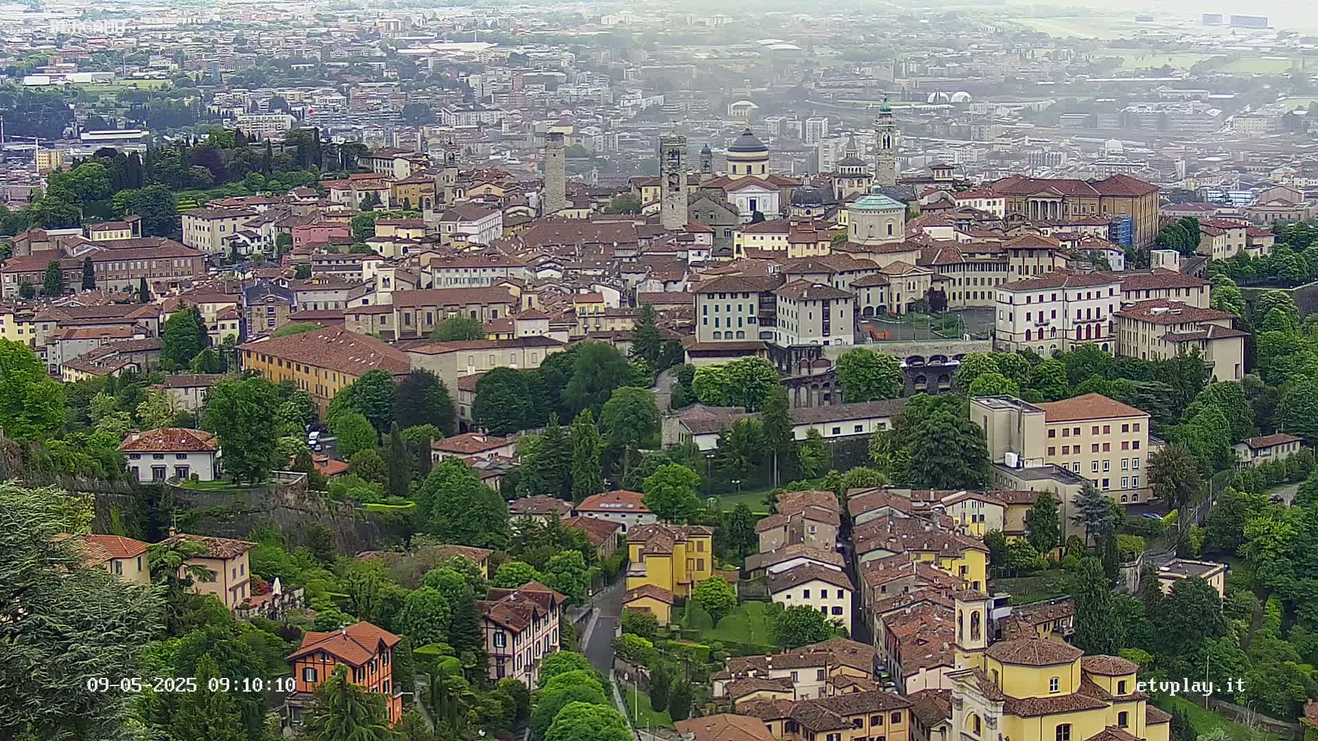 A sprawling cityscape of numerous closely clustered buildings, mostly low-rise and terracotta-roofed, nestled within abundant green foliage, under an overcast sky.