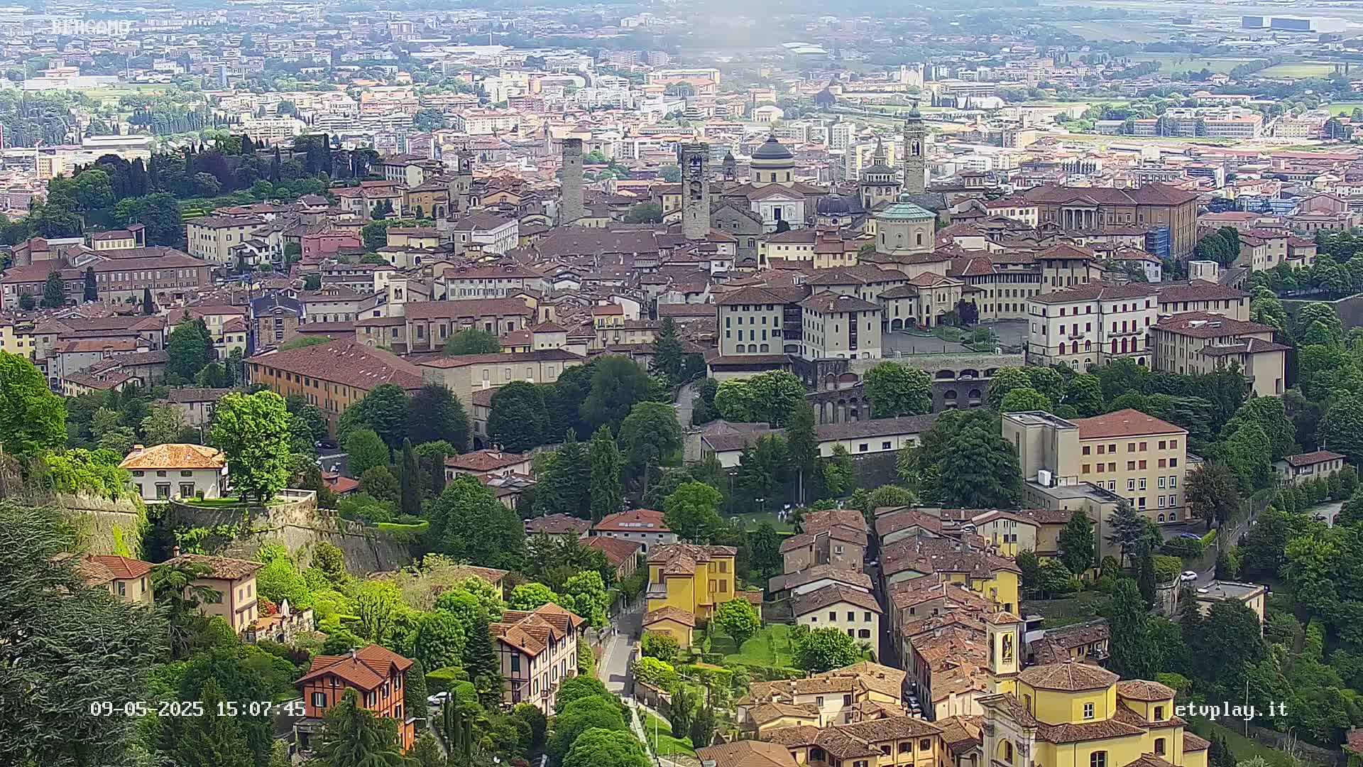 A sprawling, historic city with numerous brown-roofed buildings, surrounded by lush green trees and hills under a hazy sky.