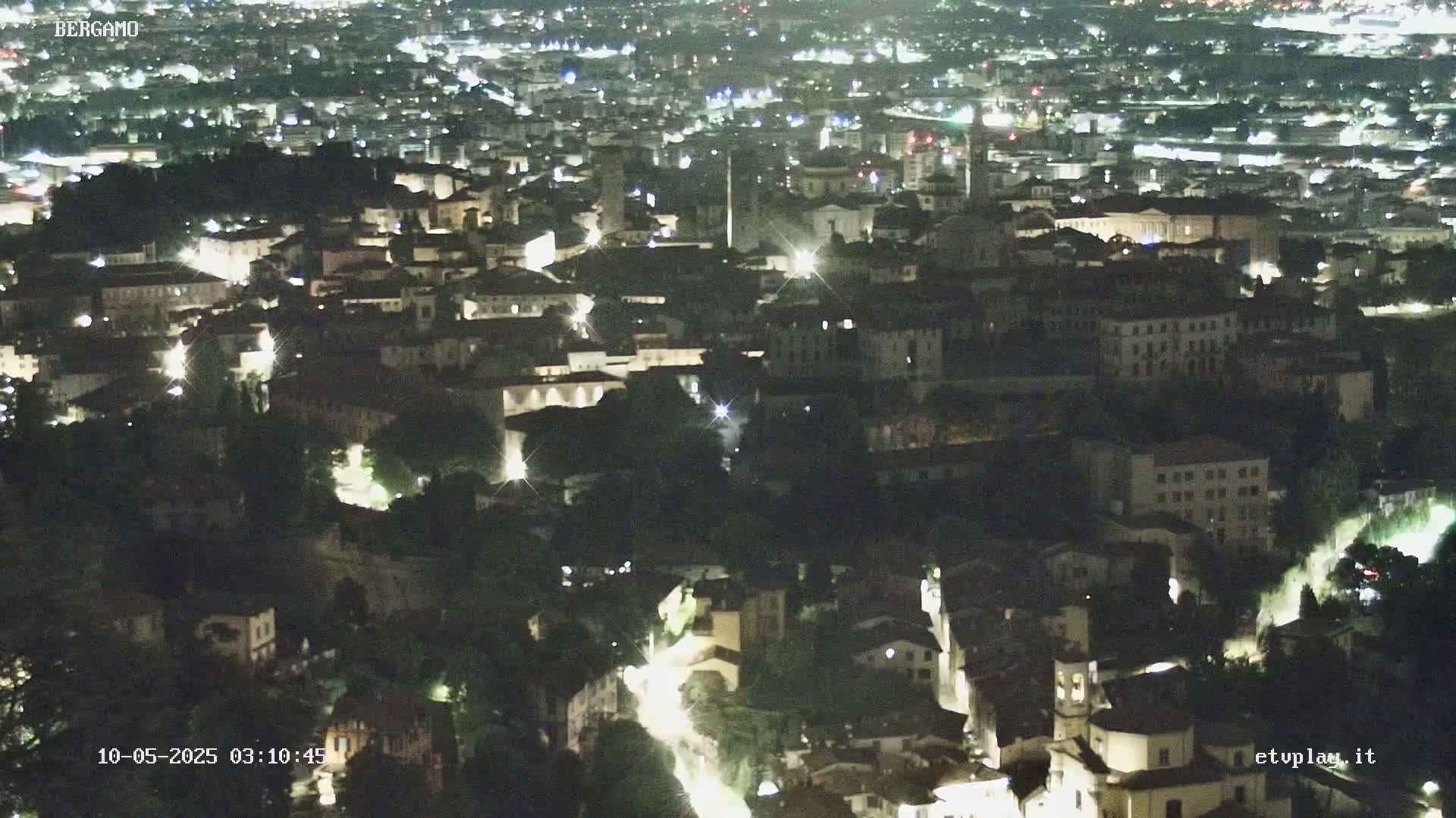 A nighttime aerial view of a city illuminated by streetlights, showing a dense collection of buildings nestled among trees.
