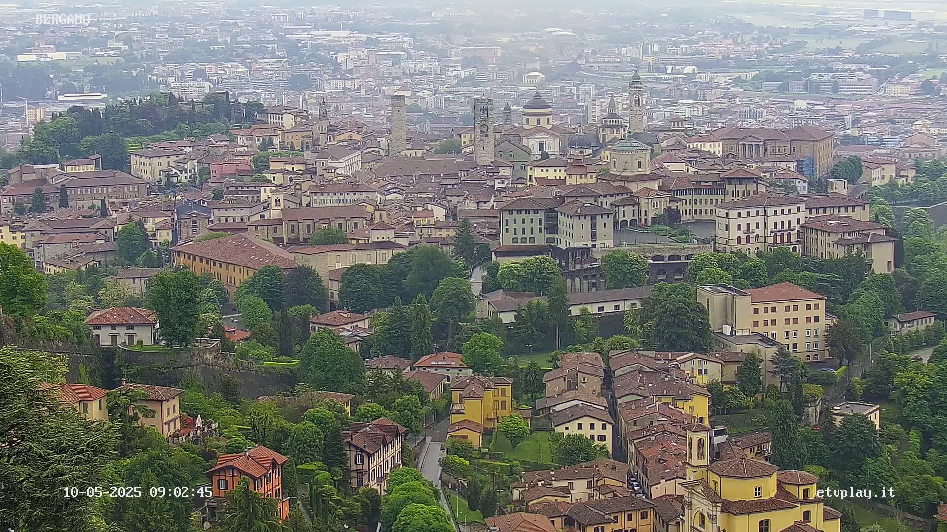 A hazy view of a European city, nestled amongst lush green hills and featuring a collection of historic buildings with terracotta roofs.
