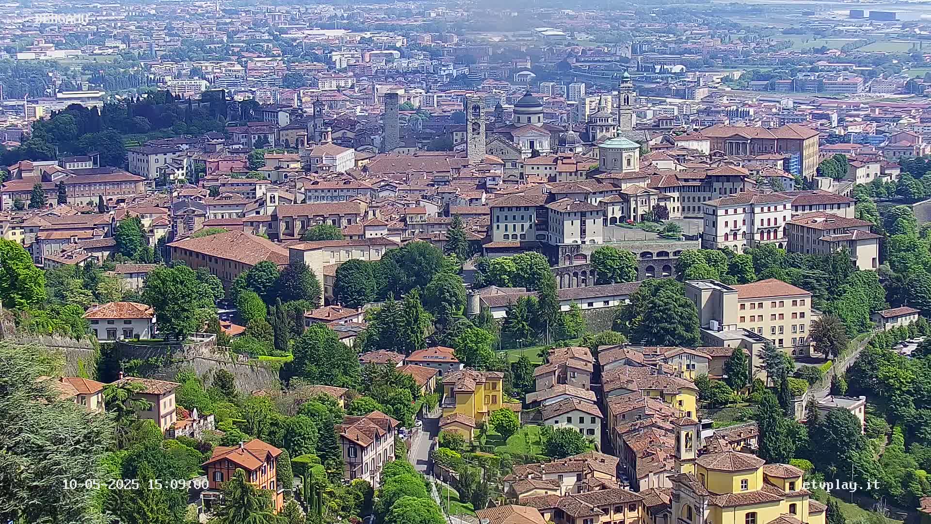 An aerial view shows a densely packed European city on a bright, sunny day, with numerous red-roofed buildings nestled amongst green trees and surrounded by a city wall.