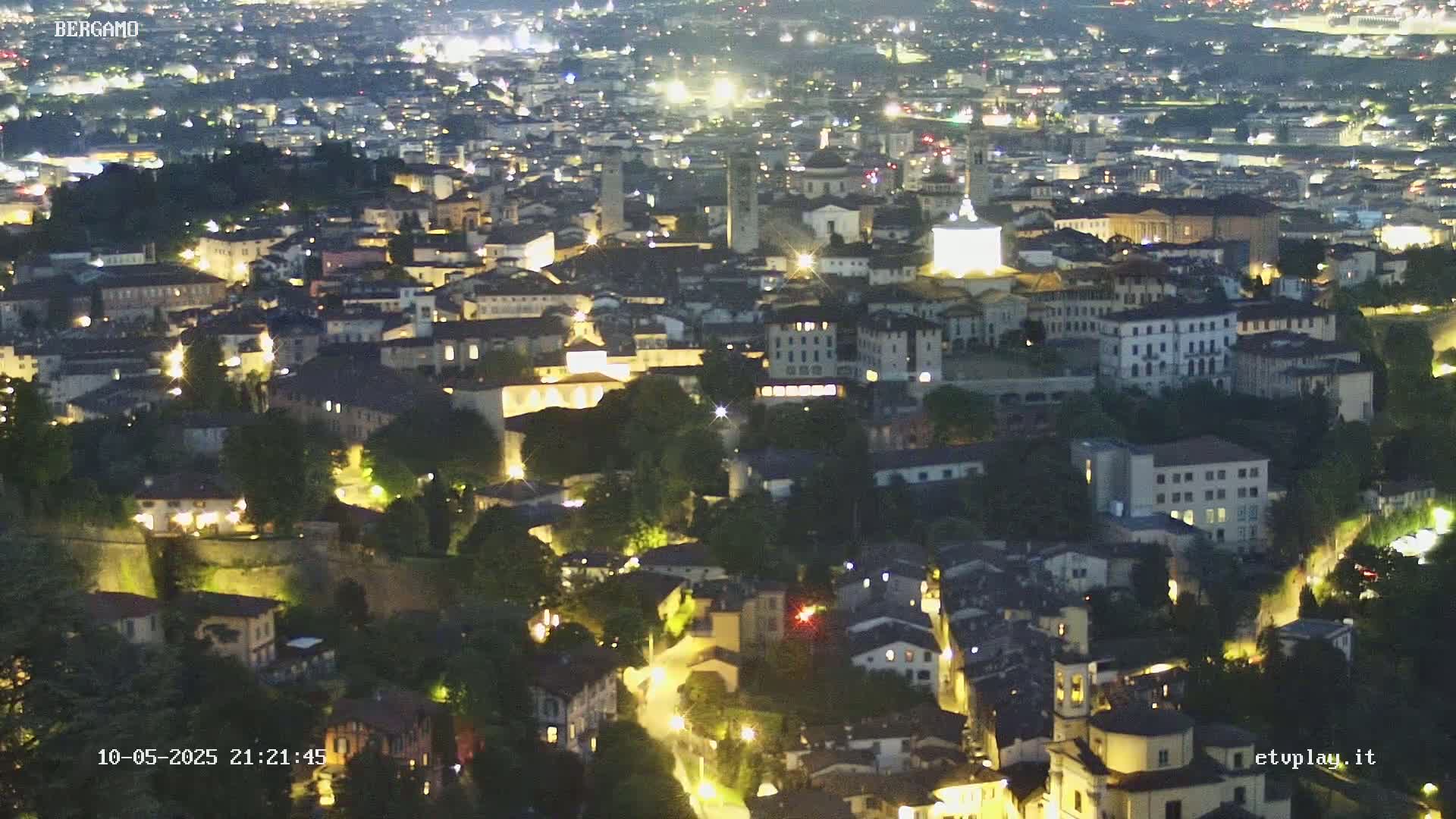 A nighttime aerial view of a city illuminated by lights, nestled amongst green hills under a clear sky.