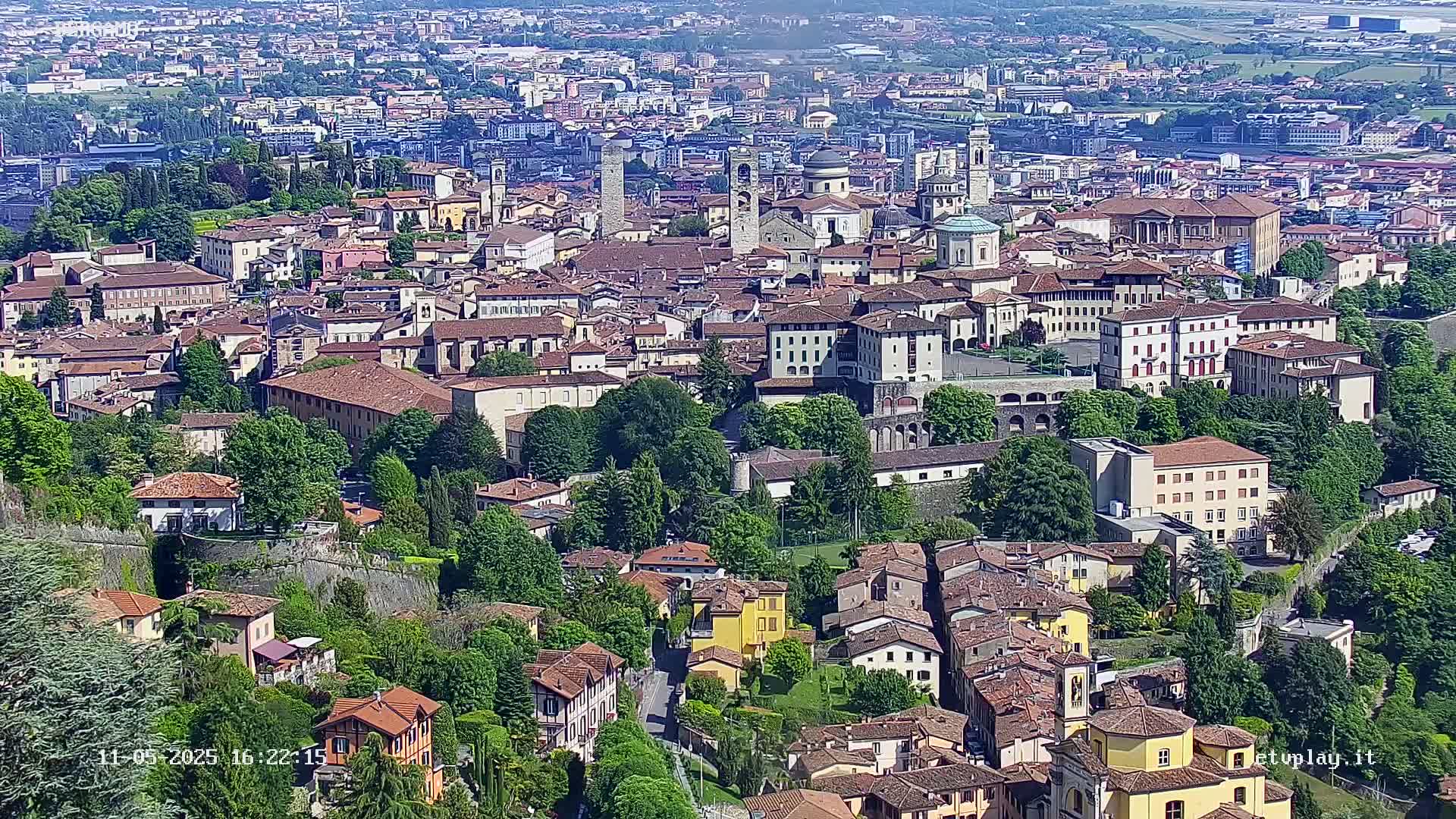 A sprawling, historic city with red-tiled roofs and lush green trees is seen from an elevated vantage point on a sunny day.