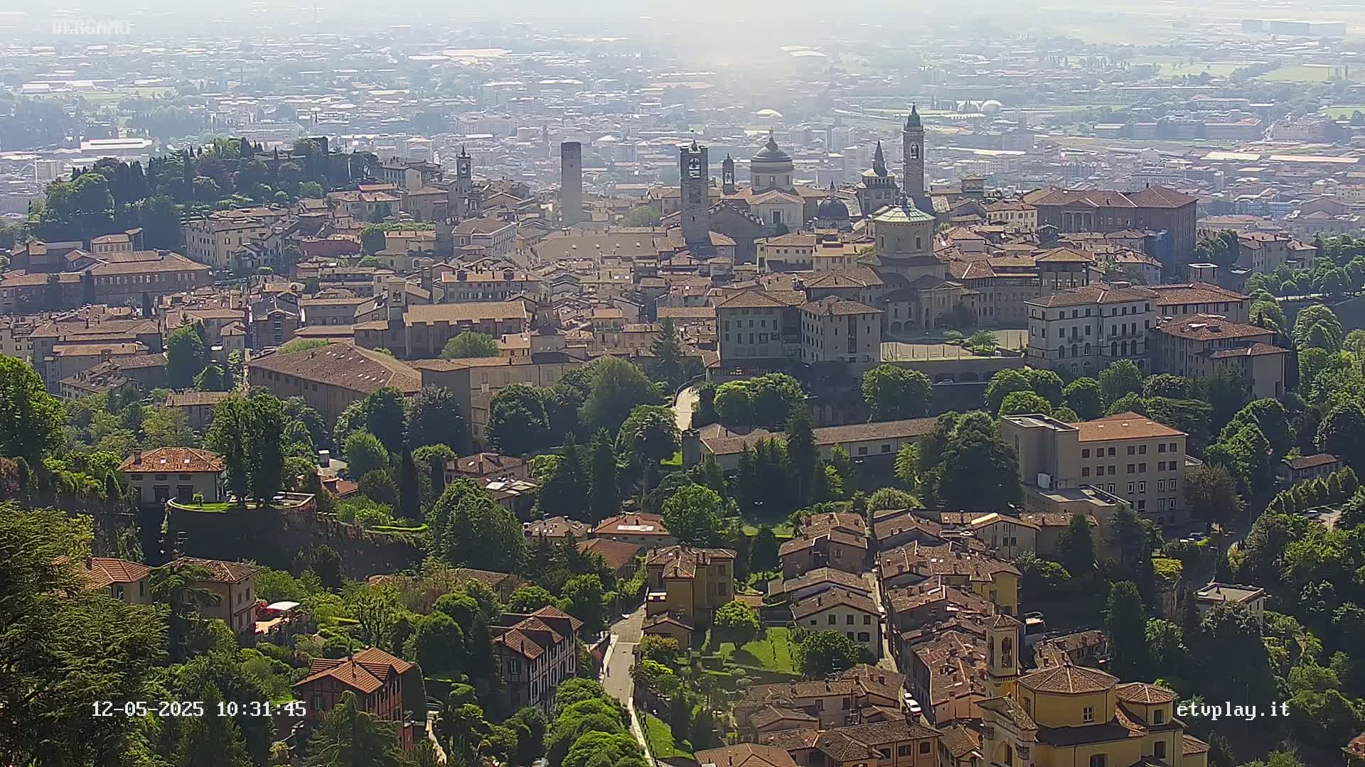 An aerial view shows a city with many buildings, churches, and green spaces on a hazy day.