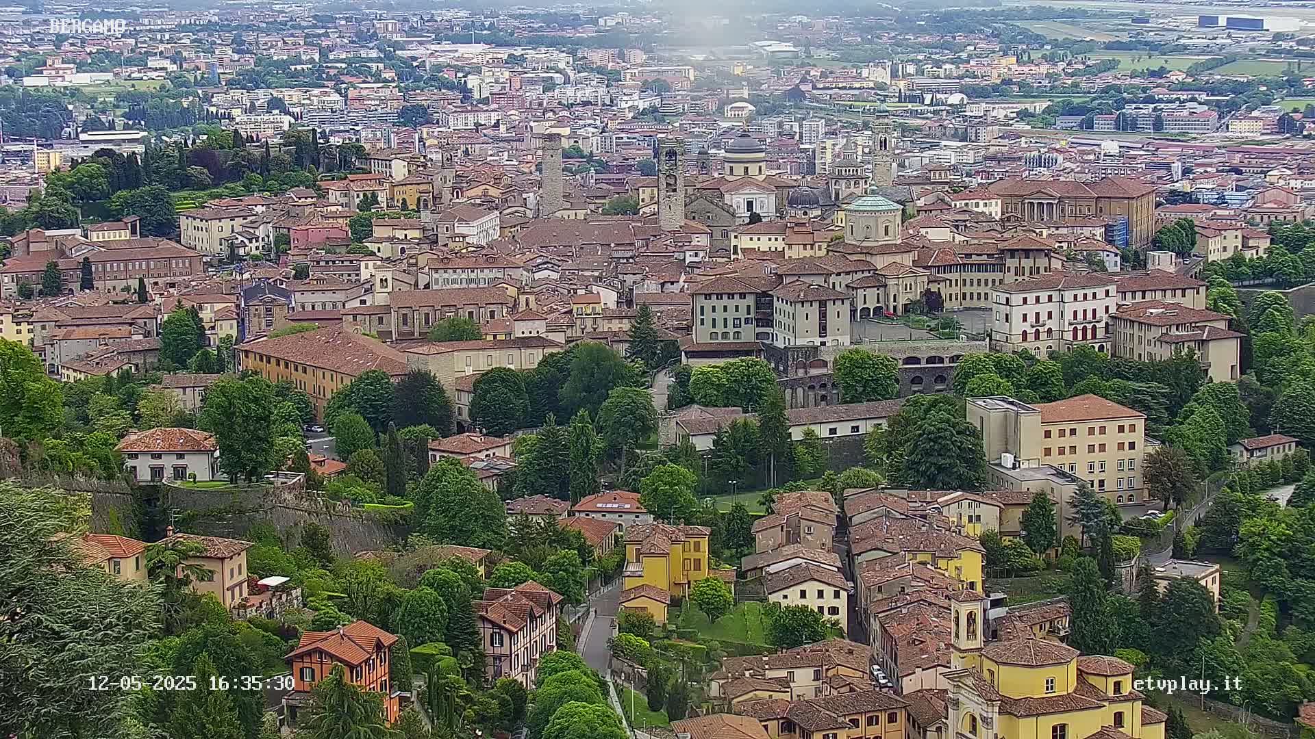 An aerial view of a city on a partly cloudy day, showing numerous closely packed buildings with red-tiled roofs surrounded by lush green trees and foliage.