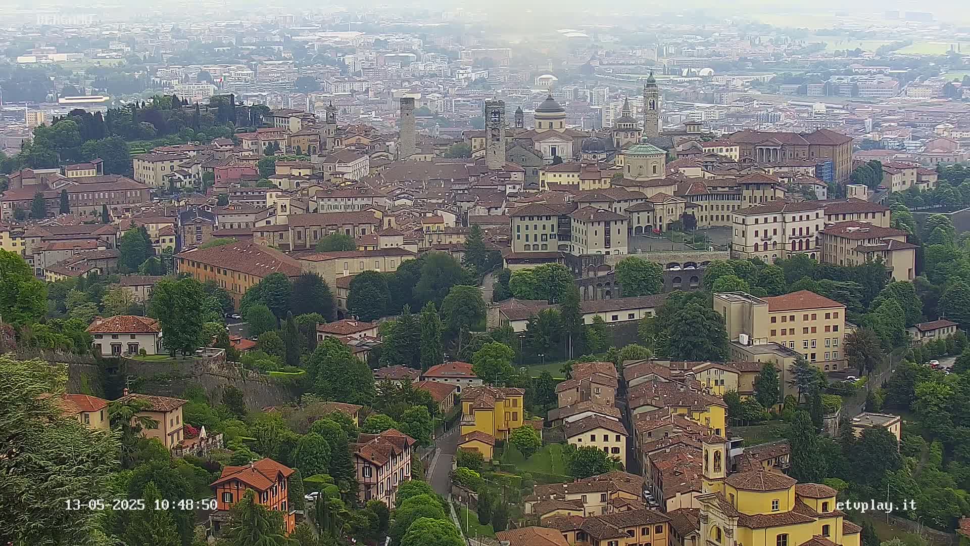 A hazy, high-angle view shows a sprawling city of many tan and brown buildings nestled among green trees and hills.
