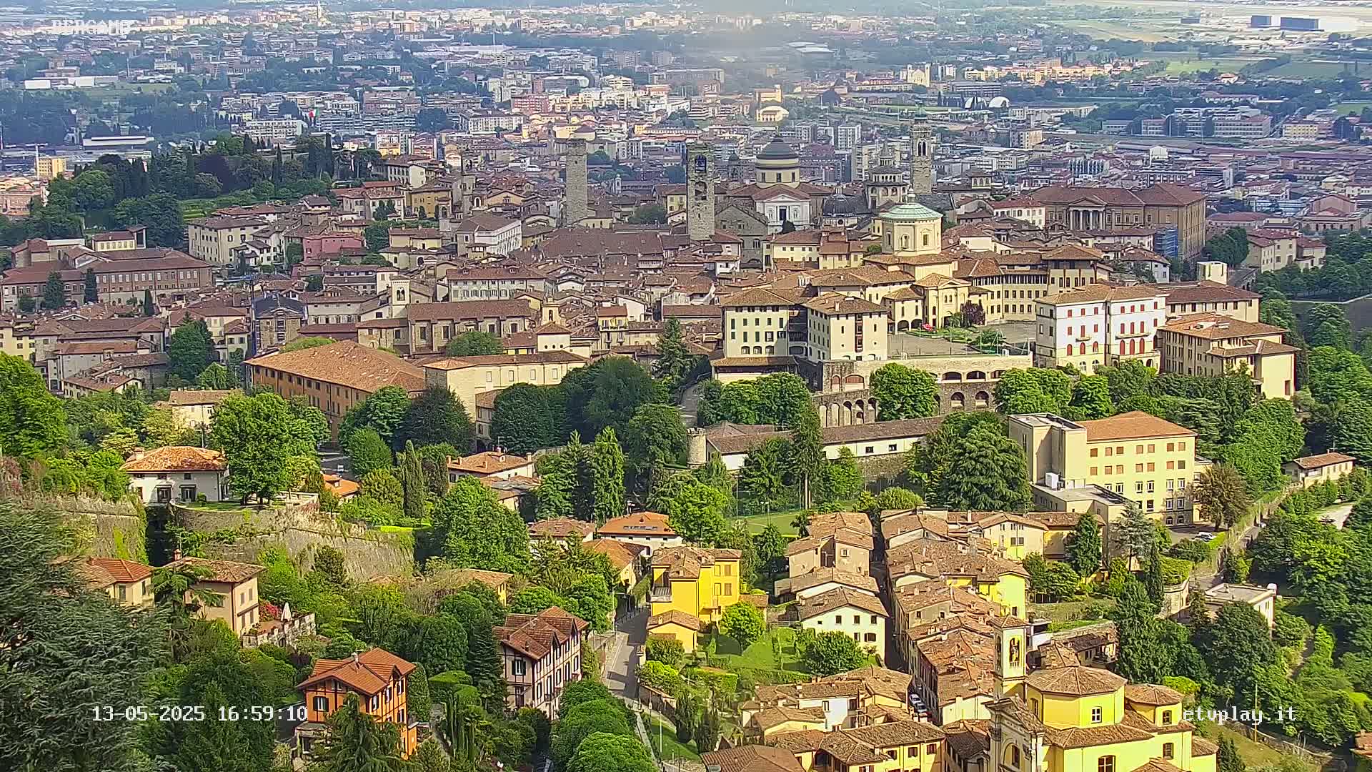 A sprawling city of tan and yellow buildings, nestled amongst lush green trees and foliage, is seen from an elevated viewpoint on a sunny day.