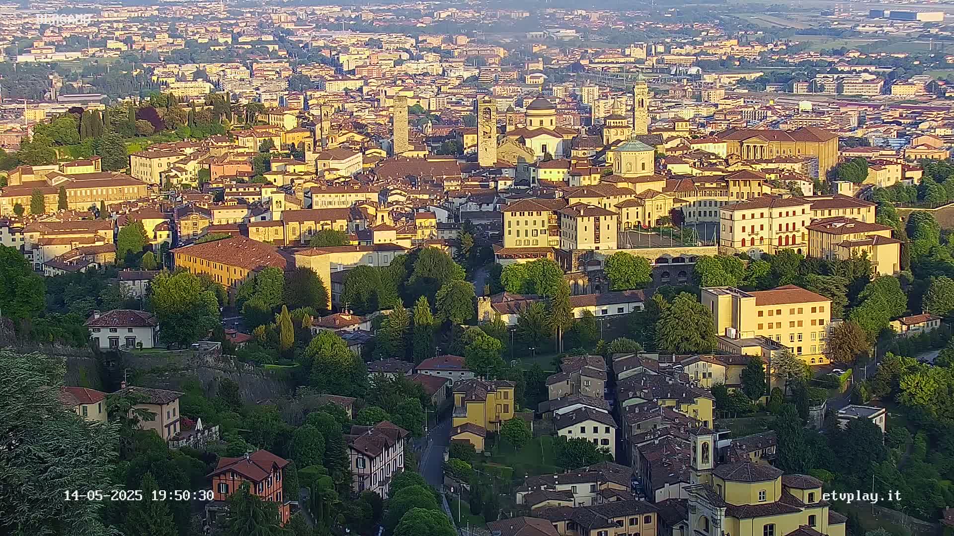 An aerial view of a densely packed European city bathed in the warm light of late afternoon sun, showing numerous buildings, trees, and a surrounding landscape.