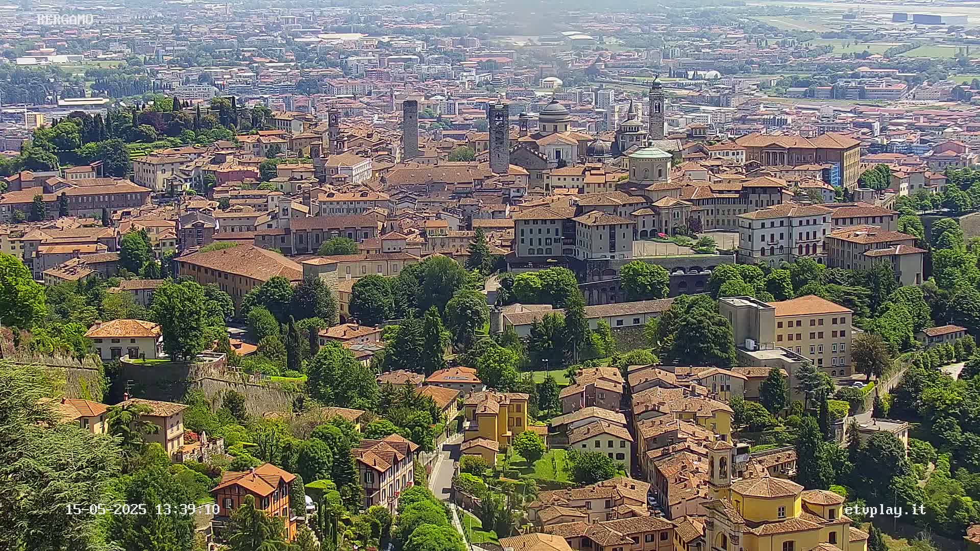 A sprawling city of terracotta-roofed buildings nestled amongst lush green trees and hills under a clear, sunny sky.