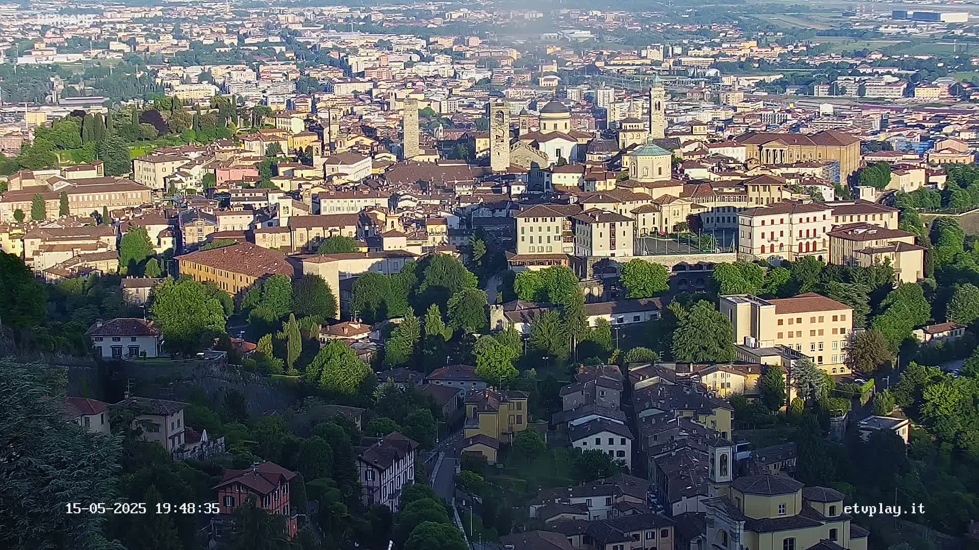 A sprawling city with many tan and brown buildings, nestled among lush green trees, is viewed from above on a sunny day.