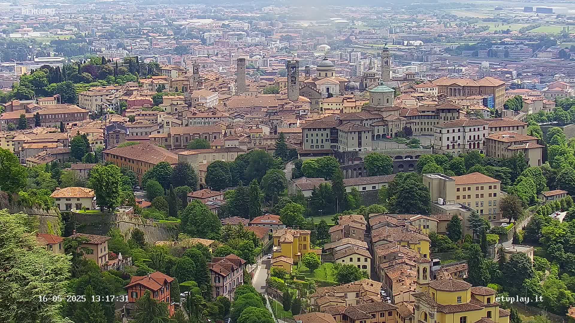An aerial view shows a sprawling city of closely clustered, terracotta-roofed buildings surrounded by lush green trees on a hazy day.