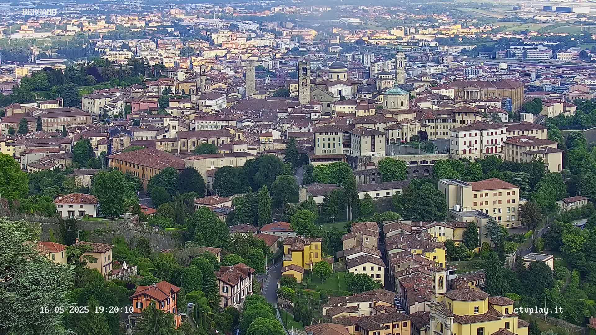 An aerial view of a densely packed European city, with many low-rise buildings and green spaces, under a partly cloudy sky.