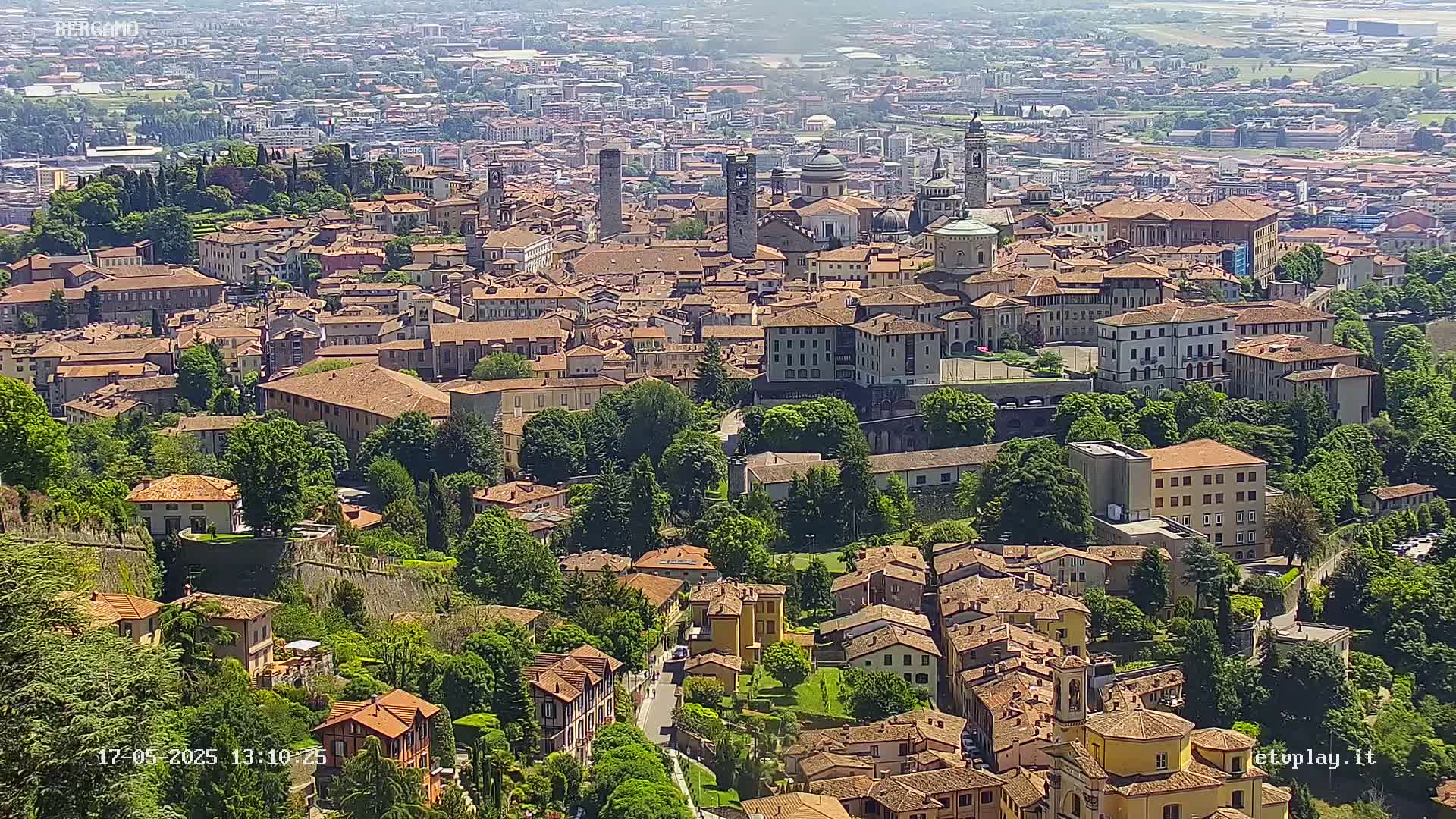 A sprawling, sunlit city of terracotta-roofed buildings nestled amongst lush green trees and hills under a clear sky.