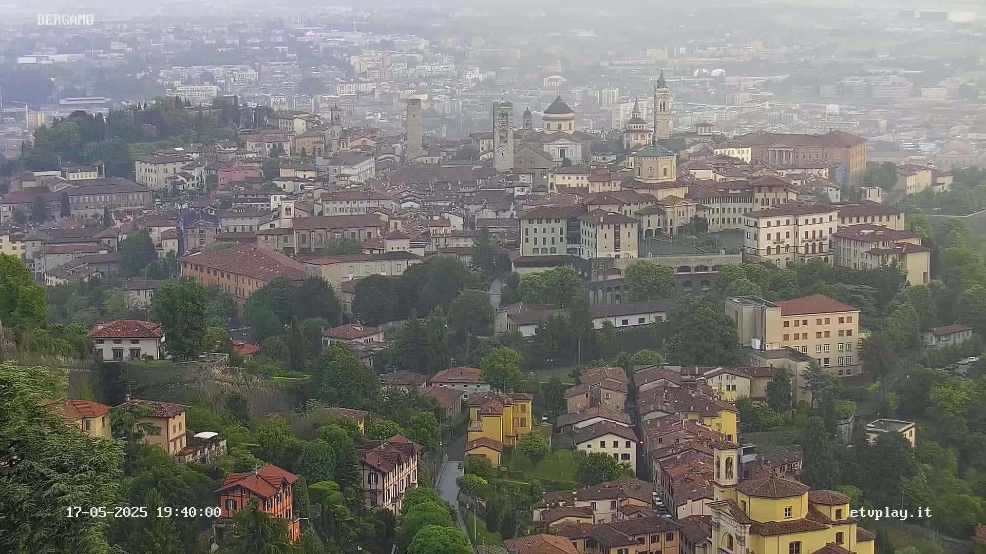 A hazy view of a sprawling city with numerous buildings, trees, and a stone wall, overlooking a lower section of town.