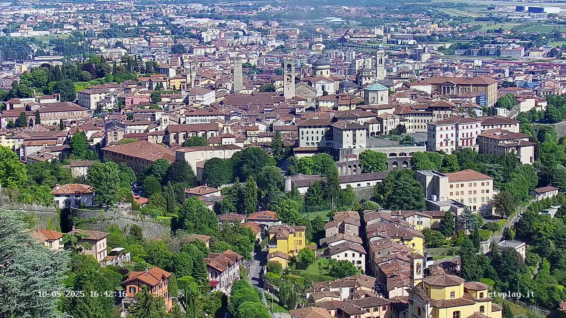A sprawling city with many closely-packed, terracotta-roofed buildings, surrounded by lush green trees and walls, under a clear sky.