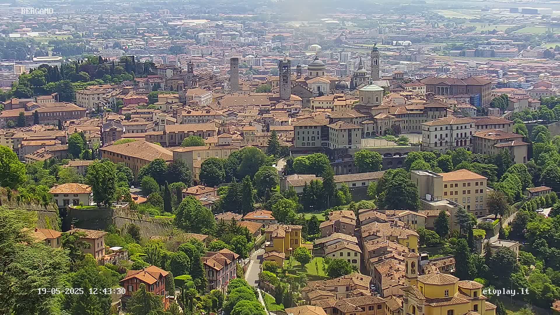 A sprawling city of tan and brown buildings, surrounded by lush green trees and hills, under a hazy sky.