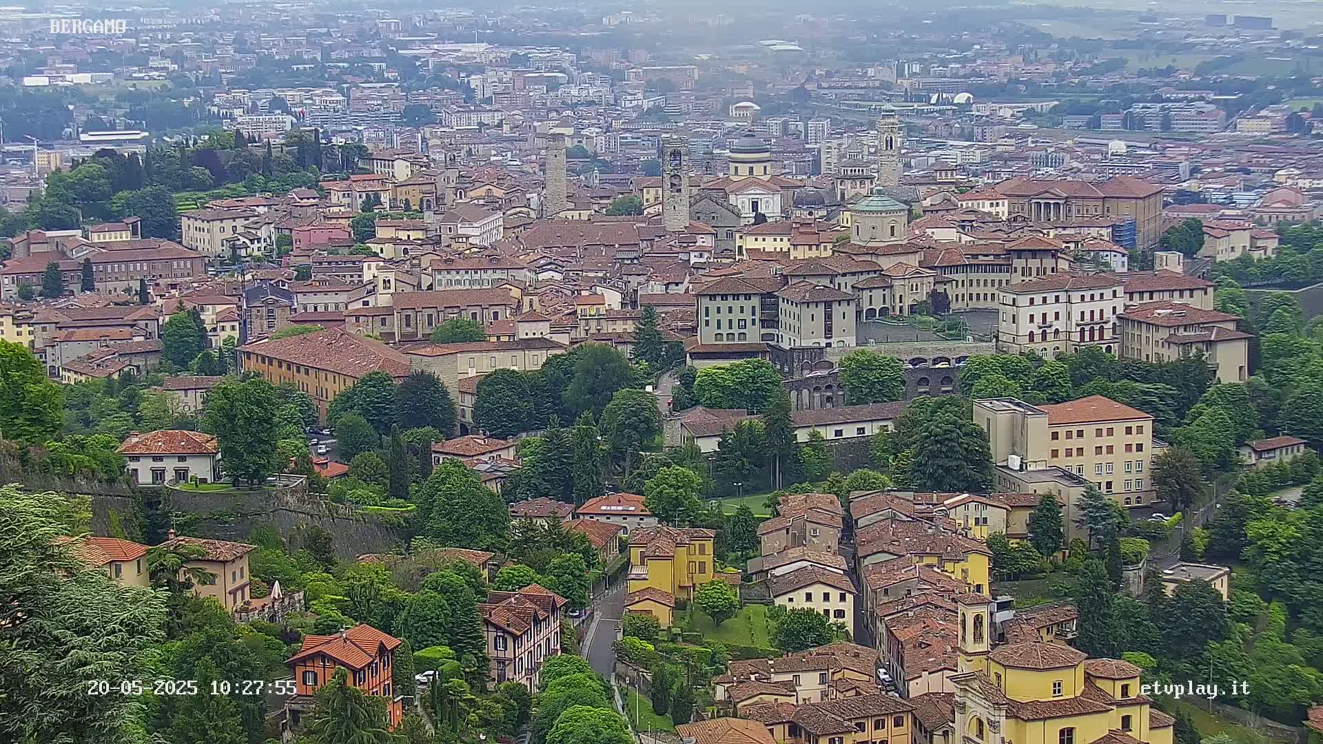 A hazy, high-angle view of a dense, historic city with numerous buildings of varying sizes and architectural styles, surrounded by lush green trees and vegetation.