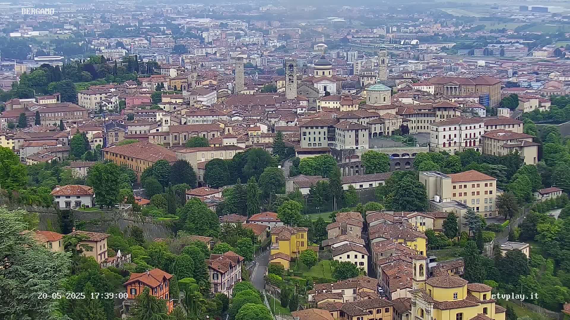 An aerial view of a dense European city on a hazy day, showing numerous buildings of varying sizes and styles nestled amongst lush green trees.