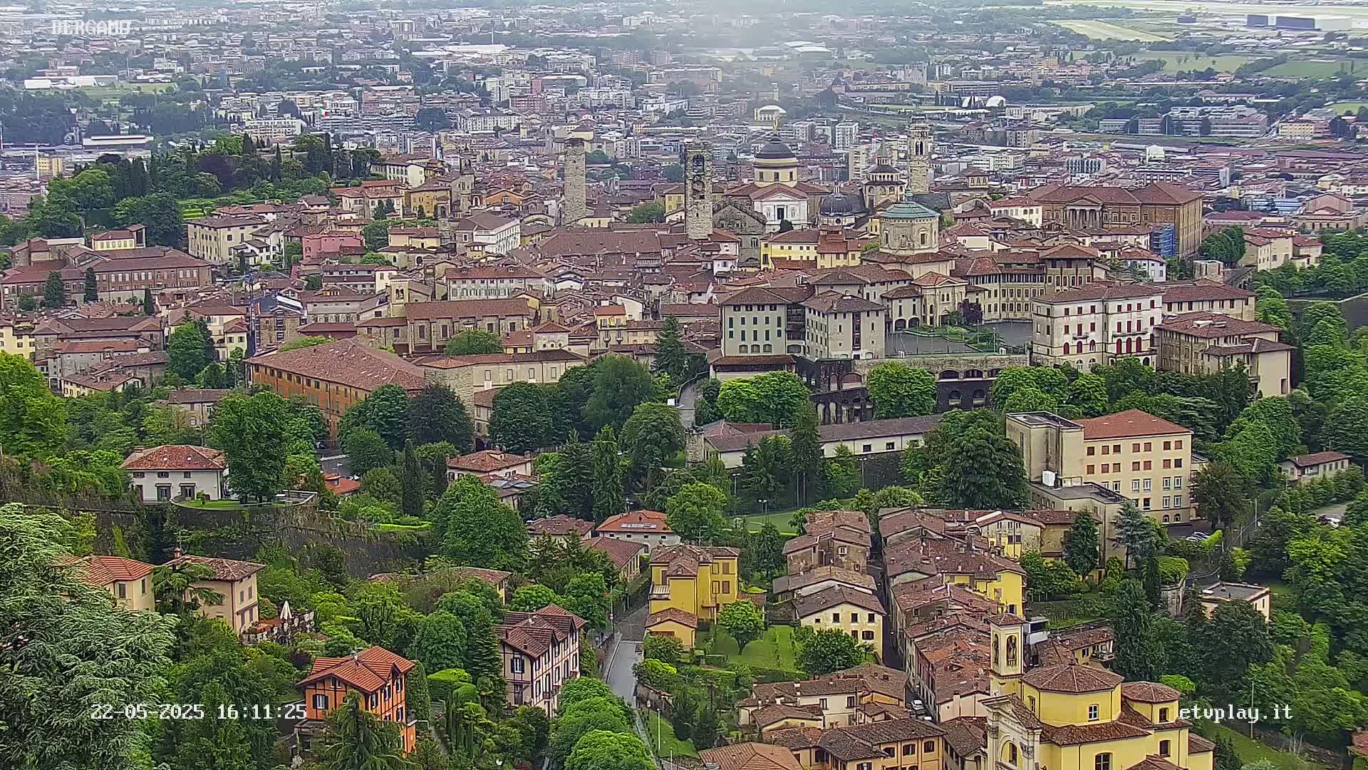 An aerial view of a dense European city on an overcast day, showcasing a mix of historic and modern buildings surrounded by lush greenery.