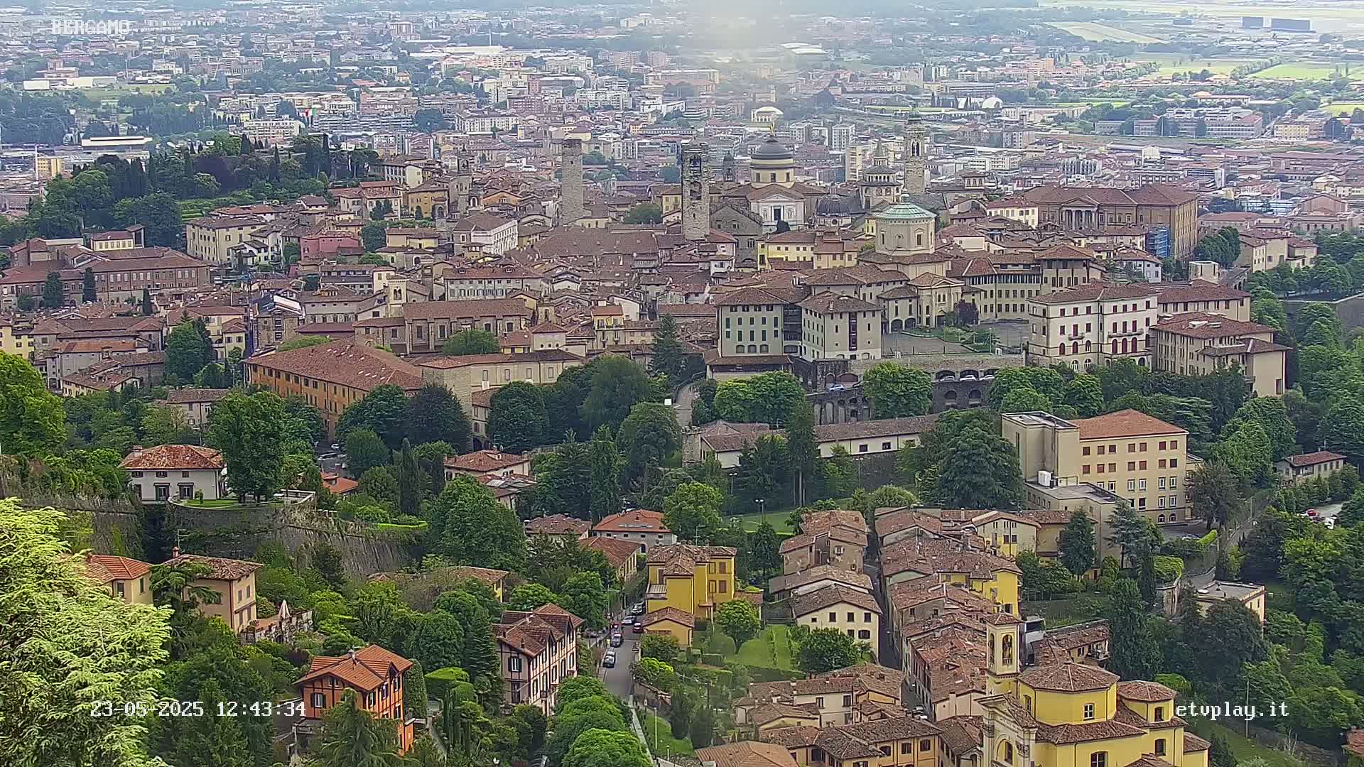 A sprawling, historic city with numerous buildings of varying heights and architectural styles, nestled amongst lush green trees under a partly cloudy sky.