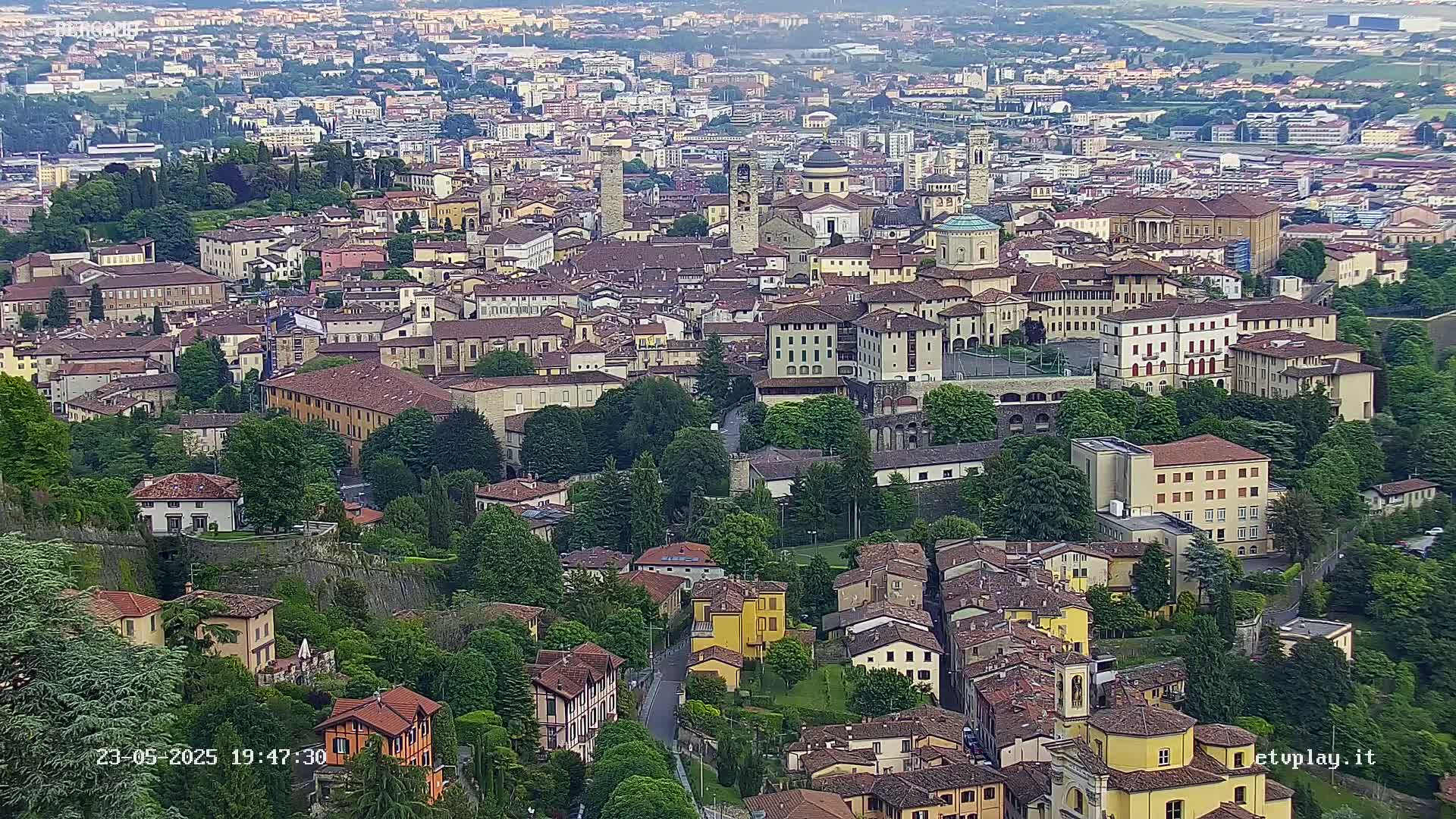 An aerial view shows a densely packed city of tan and brown buildings nestled among lush green trees and walls, under a mostly clear sky.