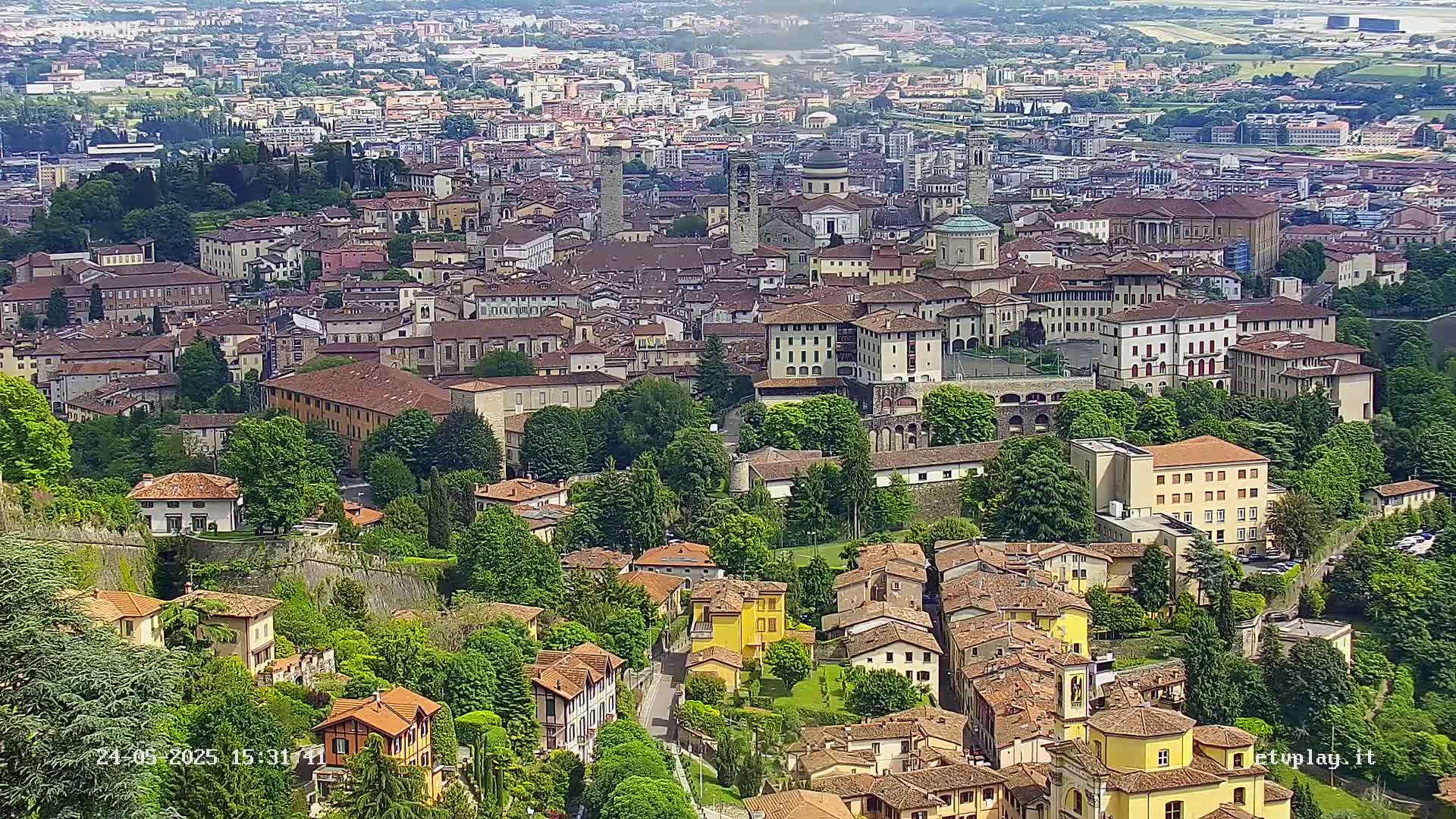 A sprawling city of terracotta-roofed buildings nestled in a lush green landscape on a partly cloudy day.