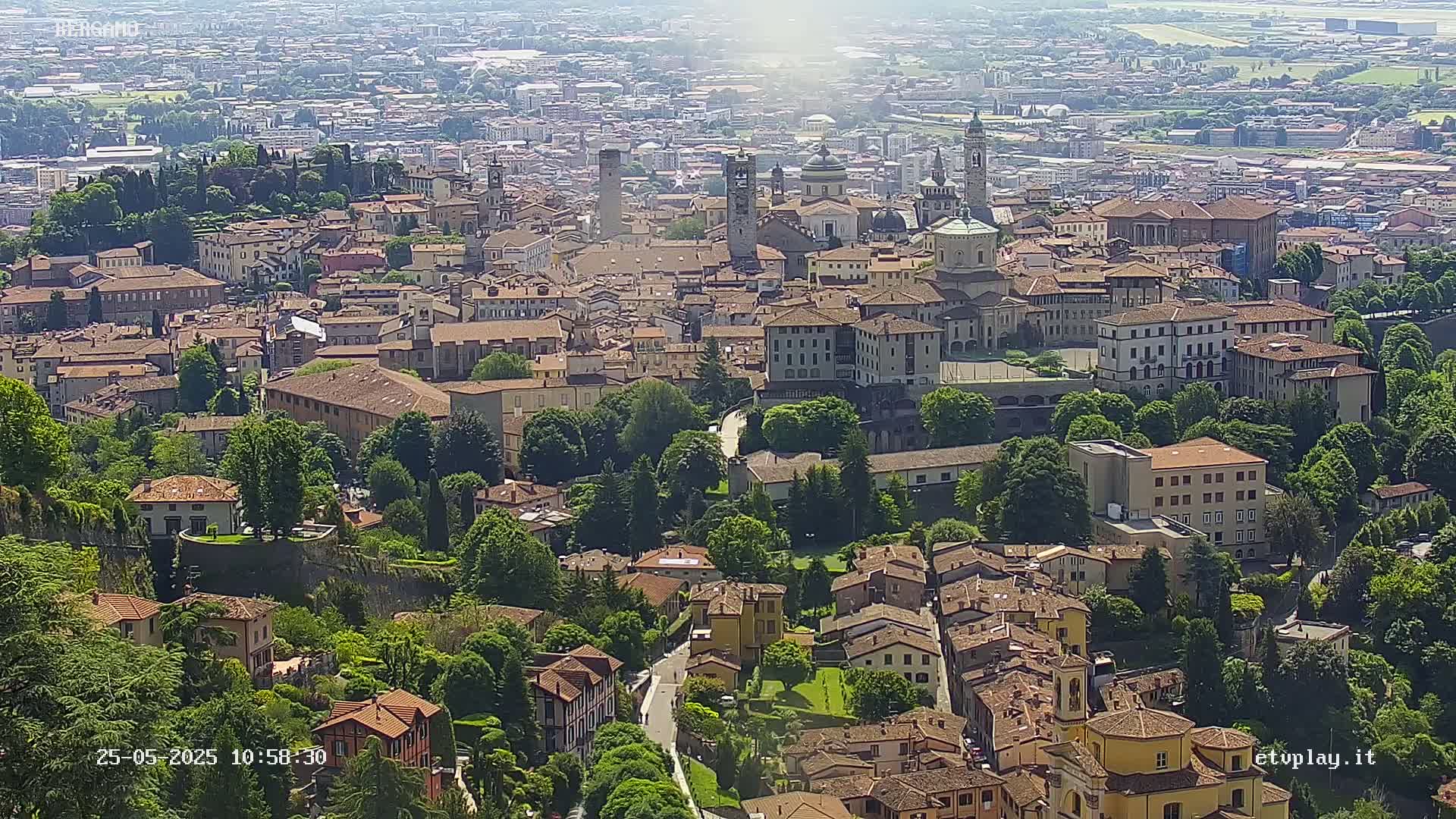 A sprawling city with numerous tan and brown buildings sits nestled amongst lush green trees and hills under a bright, sunny sky.