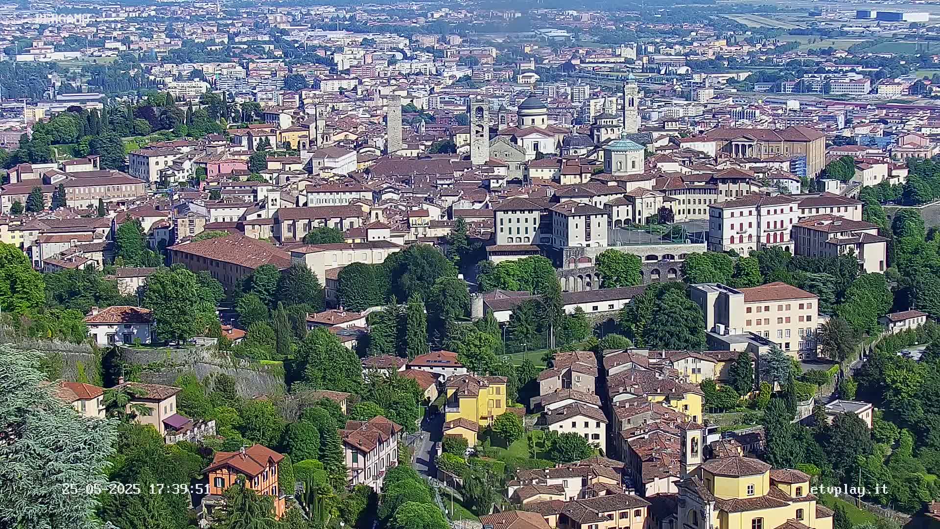 A sprawling city of closely packed buildings nestled among lush green trees and hills on a sunny day.