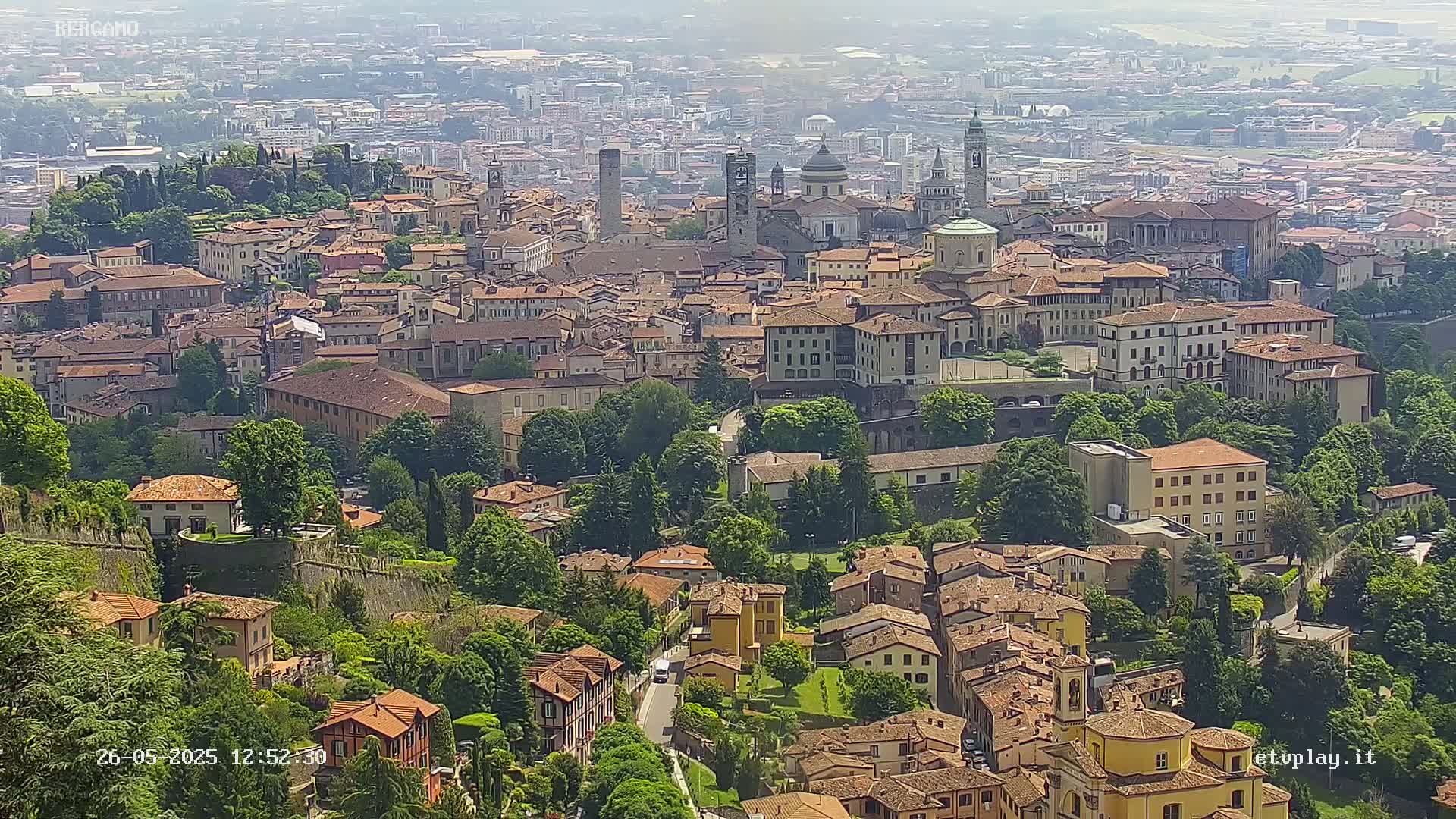 A sprawling city with terracotta-roofed buildings and green spaces is viewed from above on a hazy day.