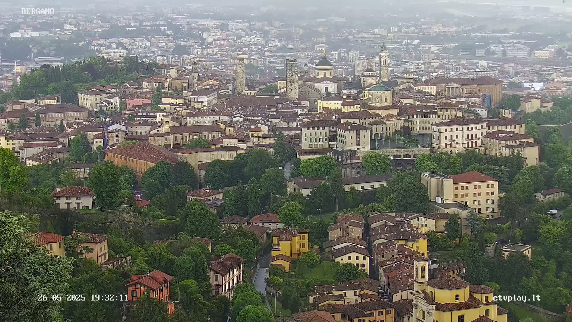 An aerial view of a city on an overcast day shows numerous buildings nestled amongst lush green trees.