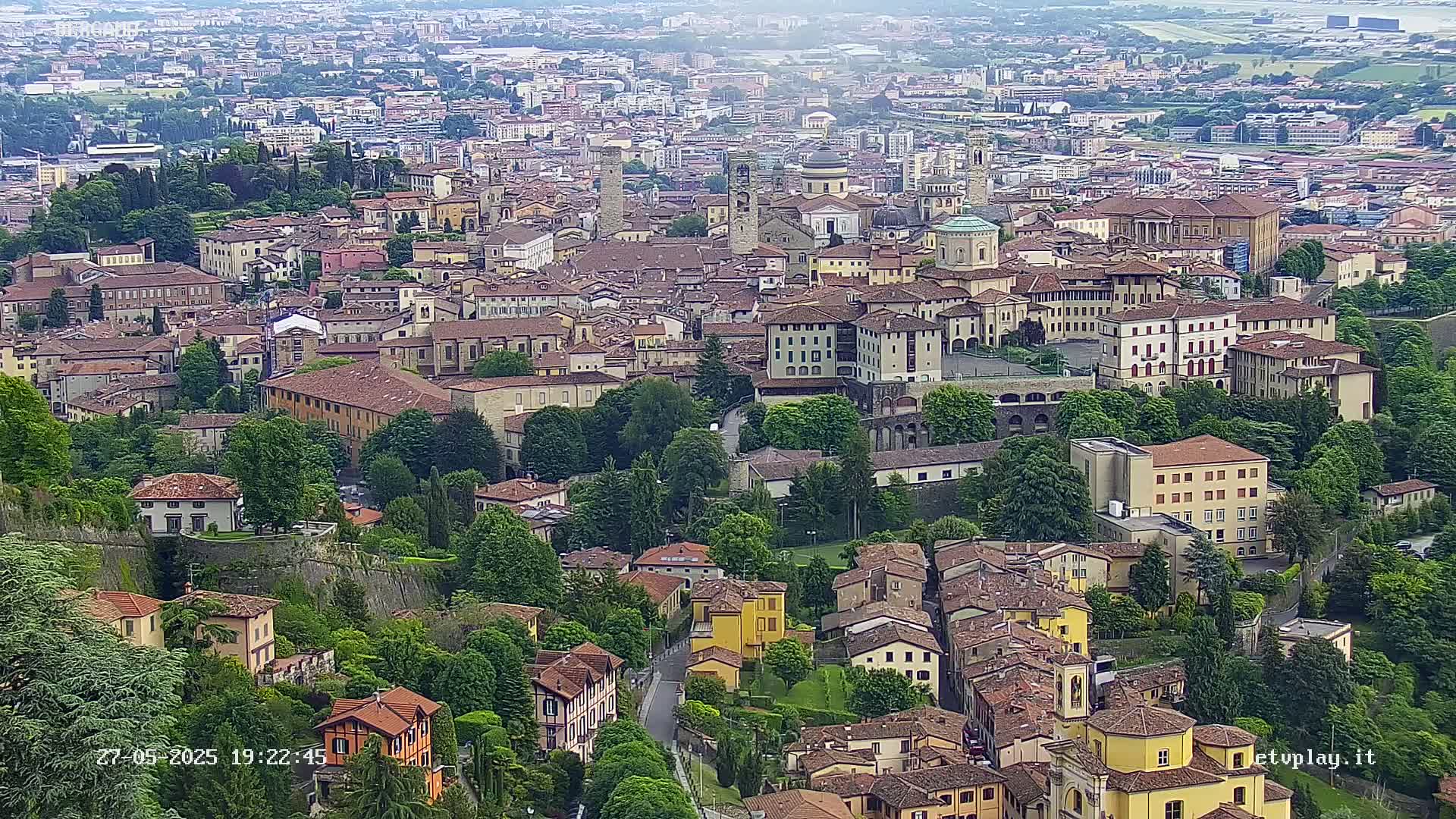 A high-angle, long shot shows a densely packed city with many brown-roofed buildings, surrounded by lush green trees and walls, under a hazy sky.
